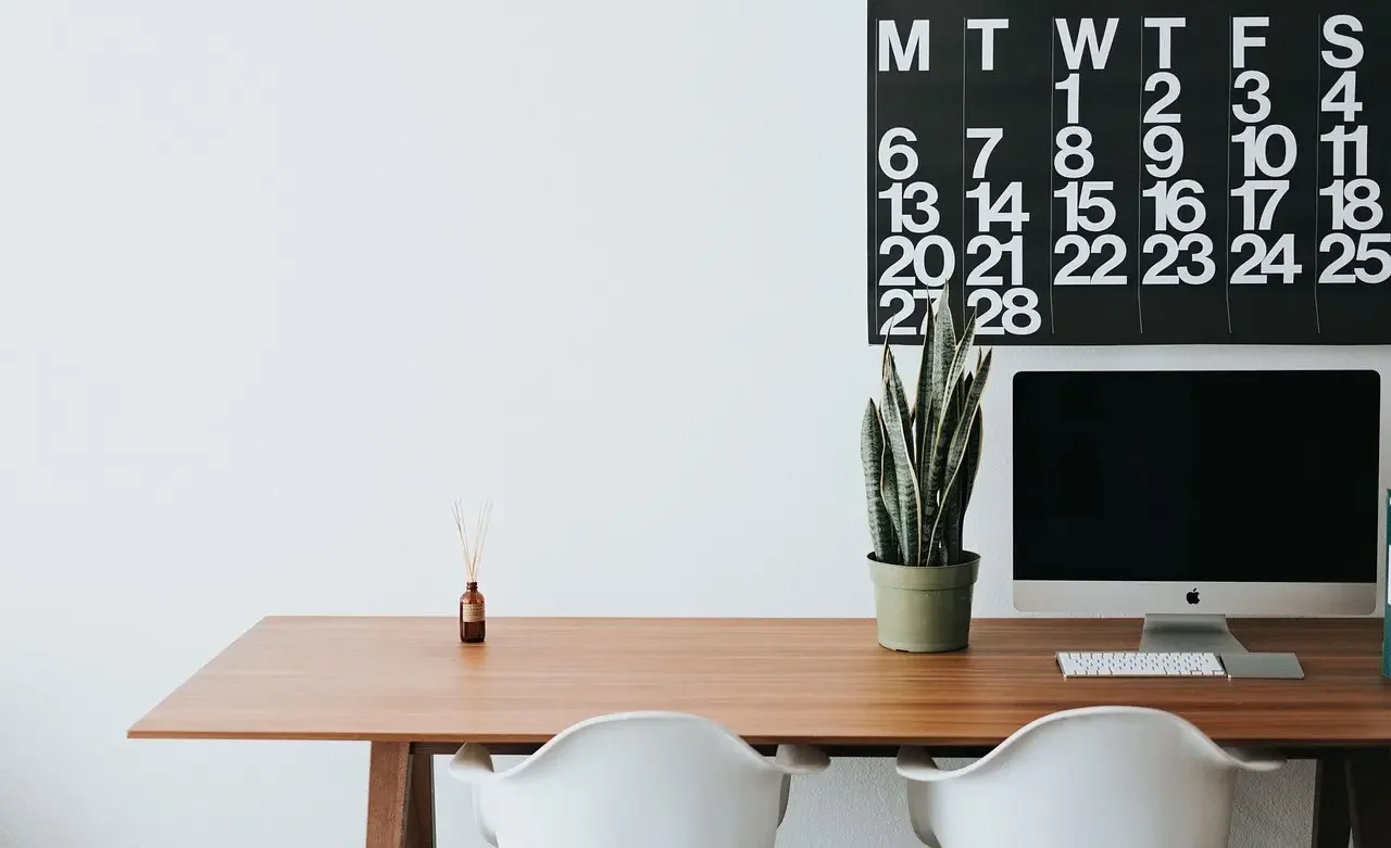 A clean, modern therapy workspace featuring a wooden desk, white chairs, a potted snake plant, and a large black and white wall calendar