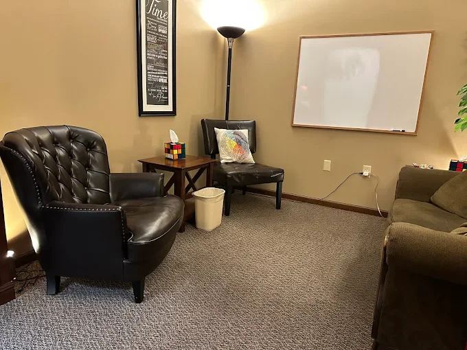 An office or waiting room with brown leather chairs, a small wooden table, a whiteboard on the wall, a floor lamp, and a beige carpeted floor.