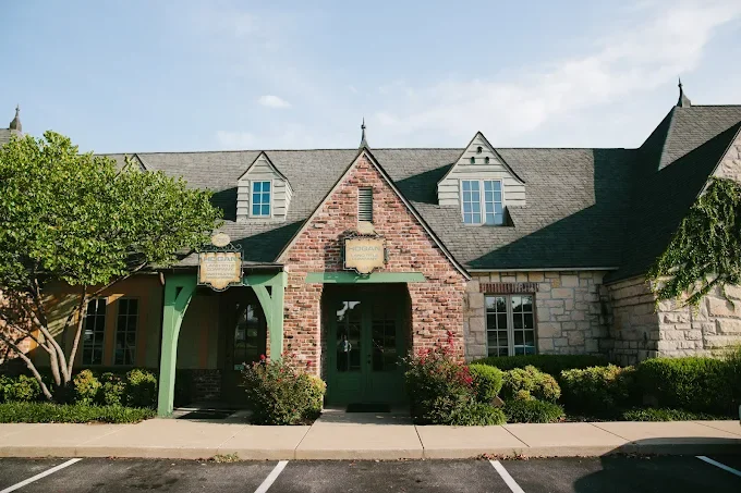 A building with a brick and stone facade, green door, and gabled roof, surrounded by shrubs and trees, under a partly cloudy sky.