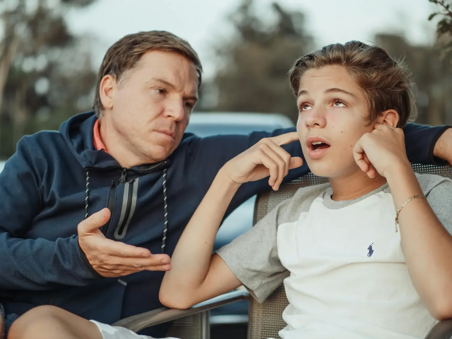 Parenting support in Springfield MO showing a father trying to communicate with a frustrated, defensive teenager who is rolling his eyes and plugging his ears.