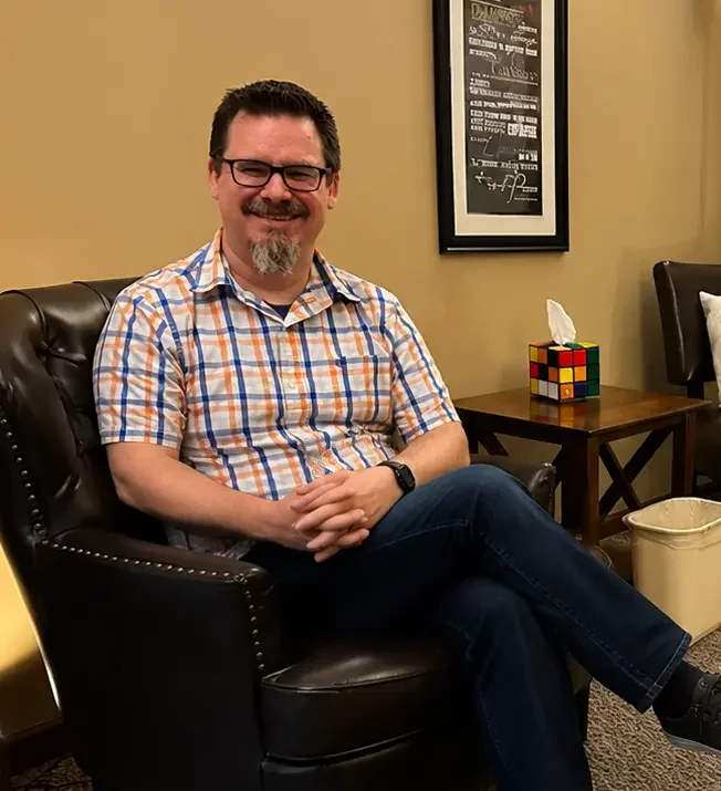 John Opalka, MA, LPC, sitting in his Springfield counseling office, smiling and wearing glasses and a plaid shirt, creating a welcoming and approachable atmosphere for clients.