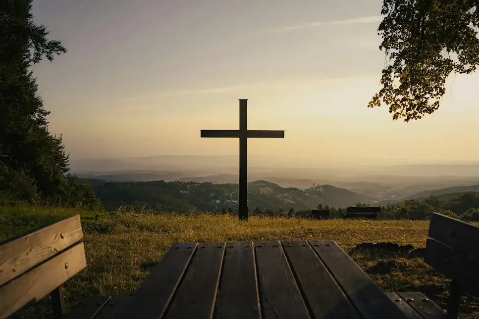 Silhouette of a Christian cross on a hill at sunset with wooden benches and a table in the foreground.