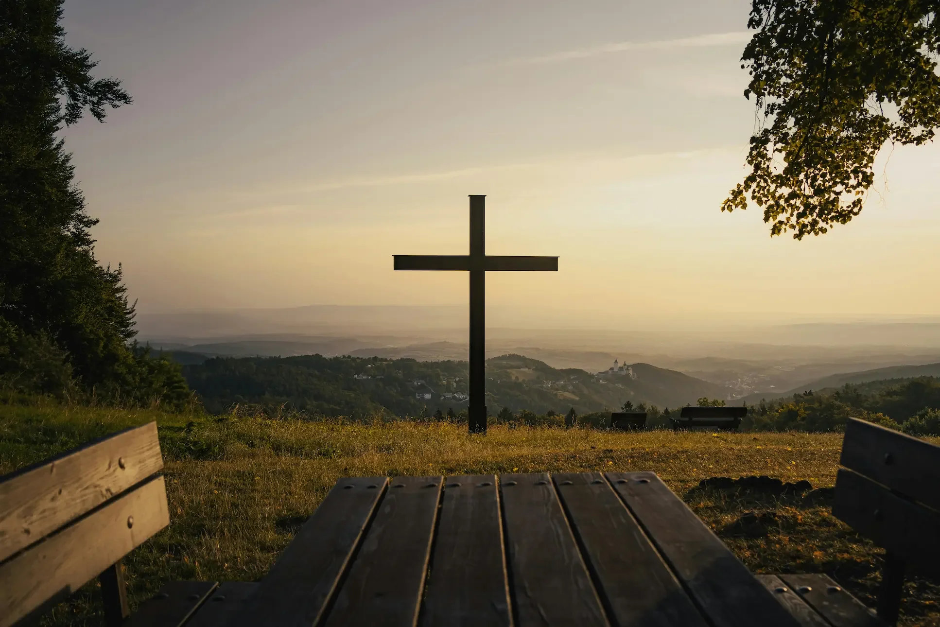 Silhouette of a Christian cross on a hill at sunset with wooden benches and a table in the foreground.