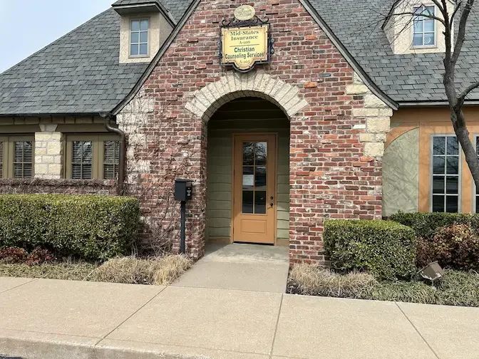 Brick building with a small arched entrance, a wooden door, and a sign above the entrance for Mid-States Insurance Christian Counseling Services, with bushes, a sidewalk, and a tree outside.