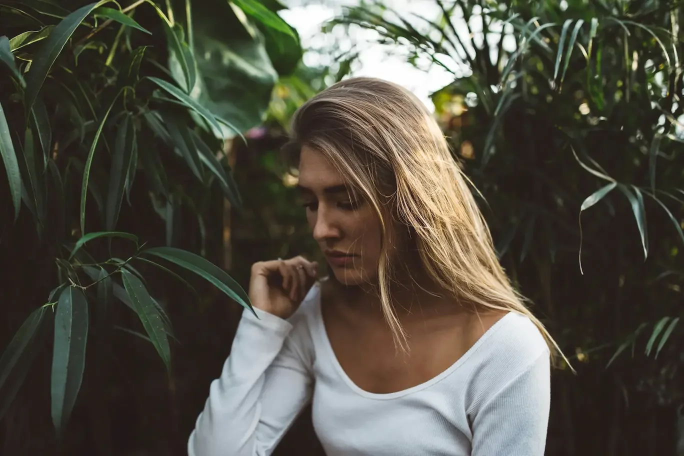 A young woman with long blonde hair looking down thoughtfully, surrounded by lush green plants, representing a moment of quiet reflection or introspection.