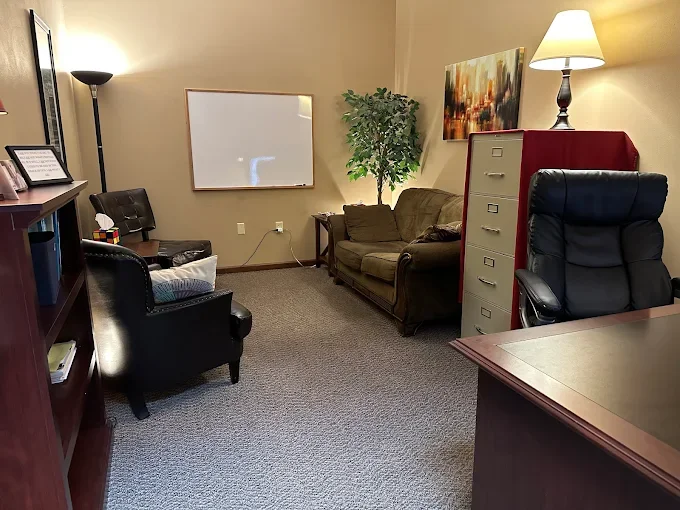 A small office or waiting room with beige walls, carpeted floor, and furniture including a black leather armchair, a sofa, a filing cabinet, a desk, a lamp, a standing floor lamp, a whiteboard, a framed picture, and a potted plant.