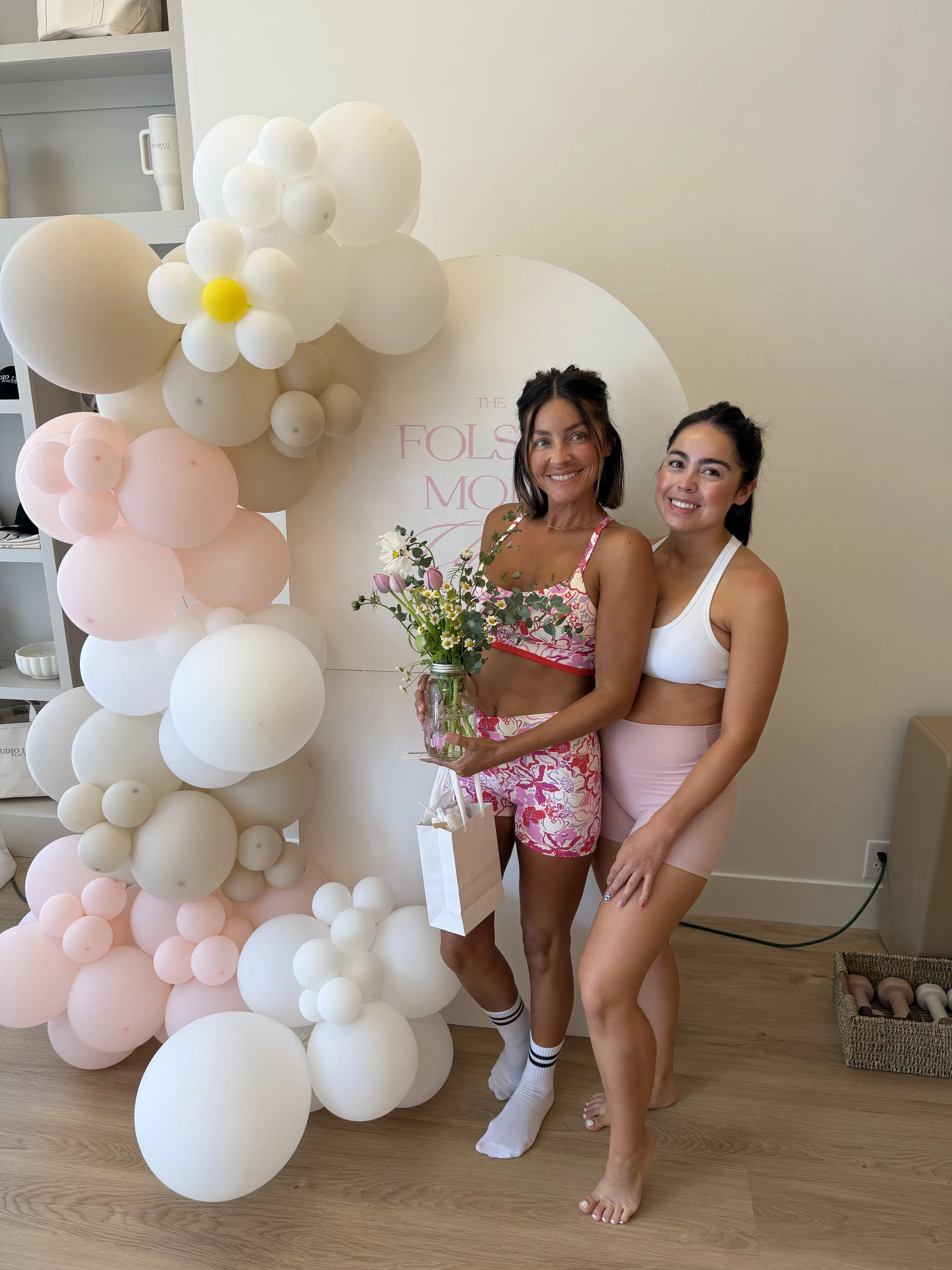 Two women standing next to a balloon display with white and pink balloons, holding a bouquet of flowers, smiling at the camera.