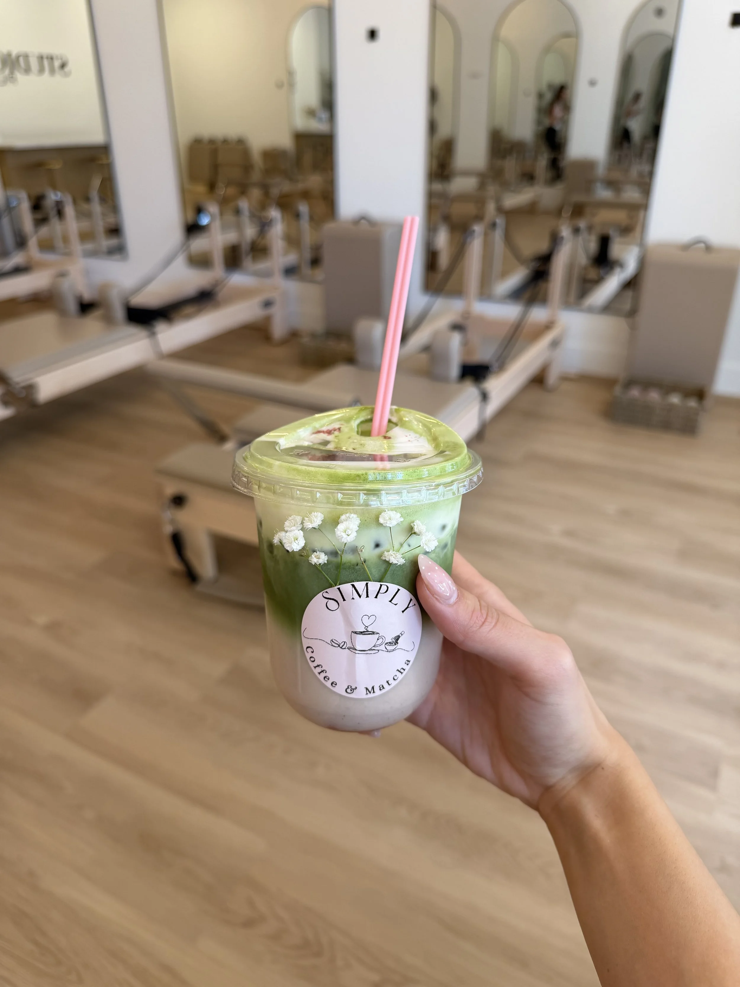 A person holding a pink and white matcha latte topped with whipped cream and small white flowers in a clear plastic cup indoors, with a blurred background of a room with wooden floors and exercise equipment.