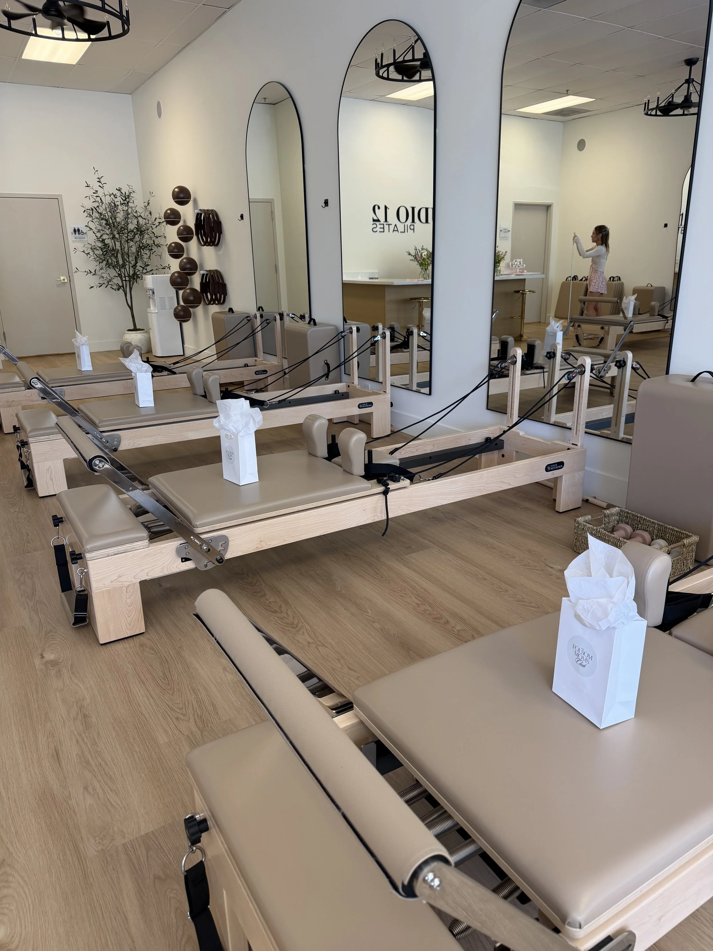 Pilates studio with multiple Reformers, large mirrors, chairs, and a woman stretching in the background.