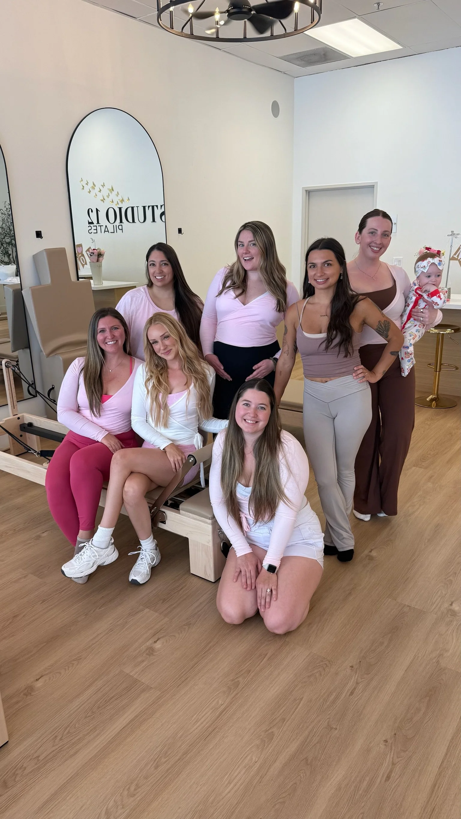 A group of nine women, some sitting and some standing, posing for a photo in what appears to be a dental or medical office. One woman is holding a baby. There are mirrors and chairs in the background.