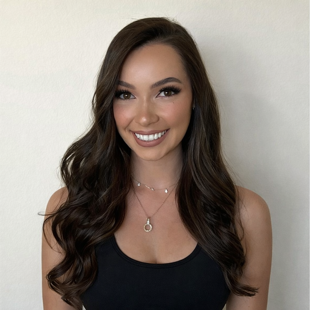 A young woman with long, wavy brown hair smiling in front of a plain white wall, wearing a black tank top and layered necklaces.