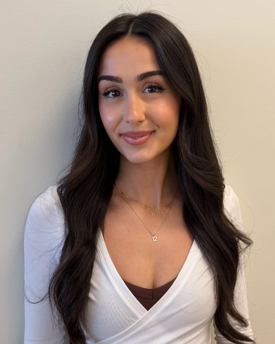 Young woman with long dark hair wearing a white top and a necklace, smiling softly in front of a plain wall.