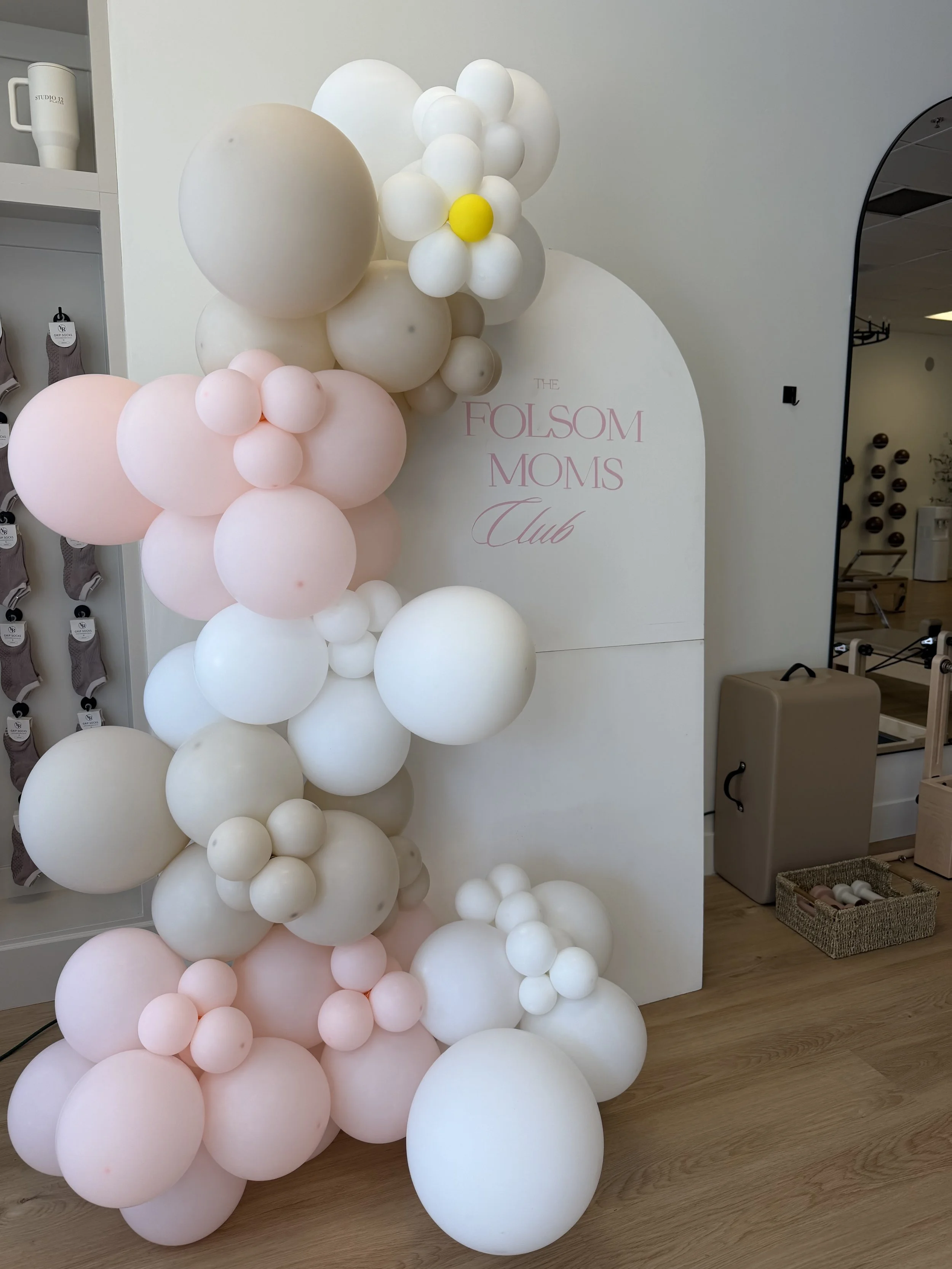 A decorative display of pink and white balloons arranged next to a sign for The Folsom Moms Club, with store shelves and a mirror visible in the background.