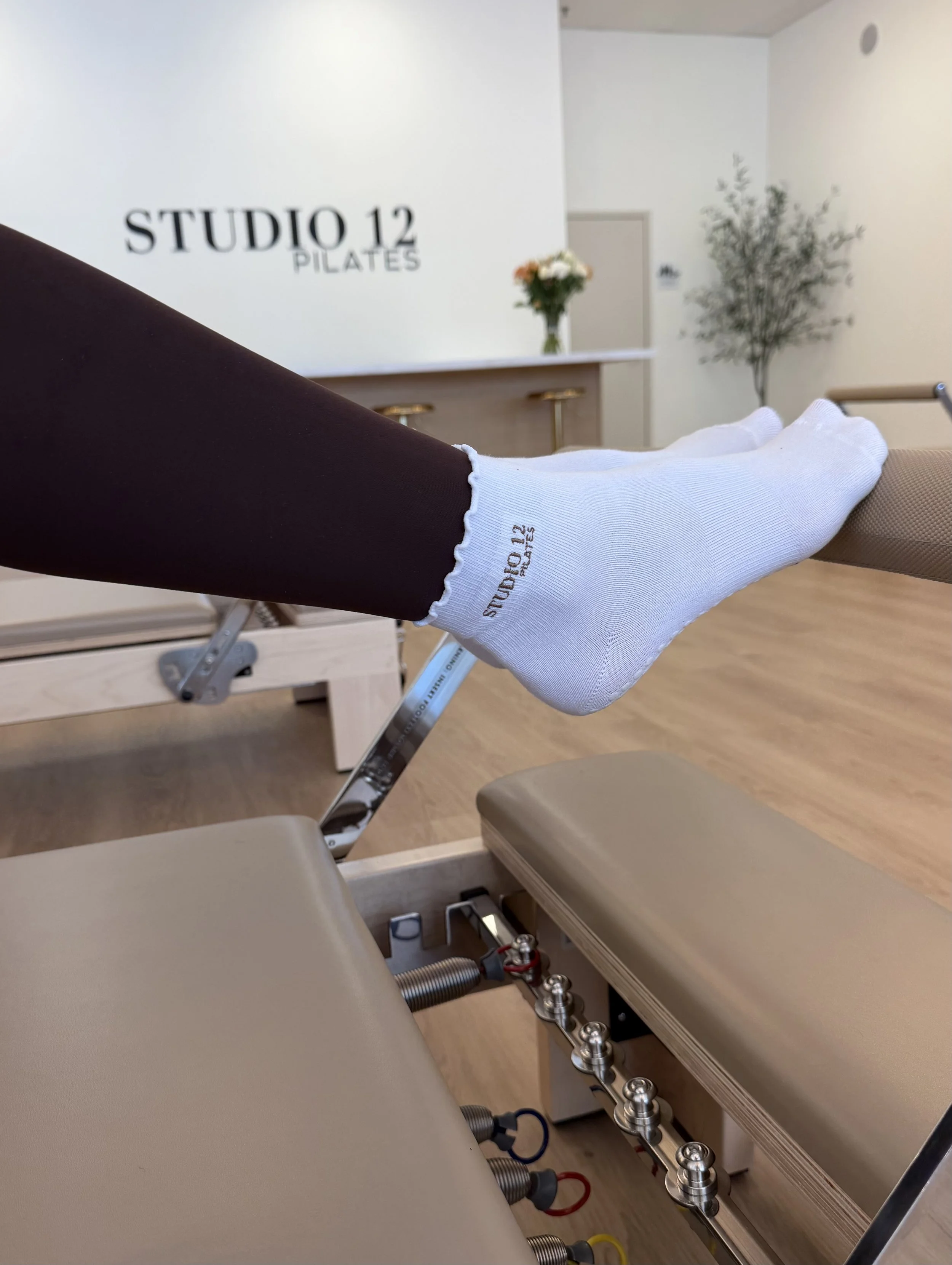 A person practicing Pilates with their feet on the reformer machine, wearing white socks with 'STUDIO 12 PILATES' written on them. The background shows a floral arrangement and the studio's logo on the wall.