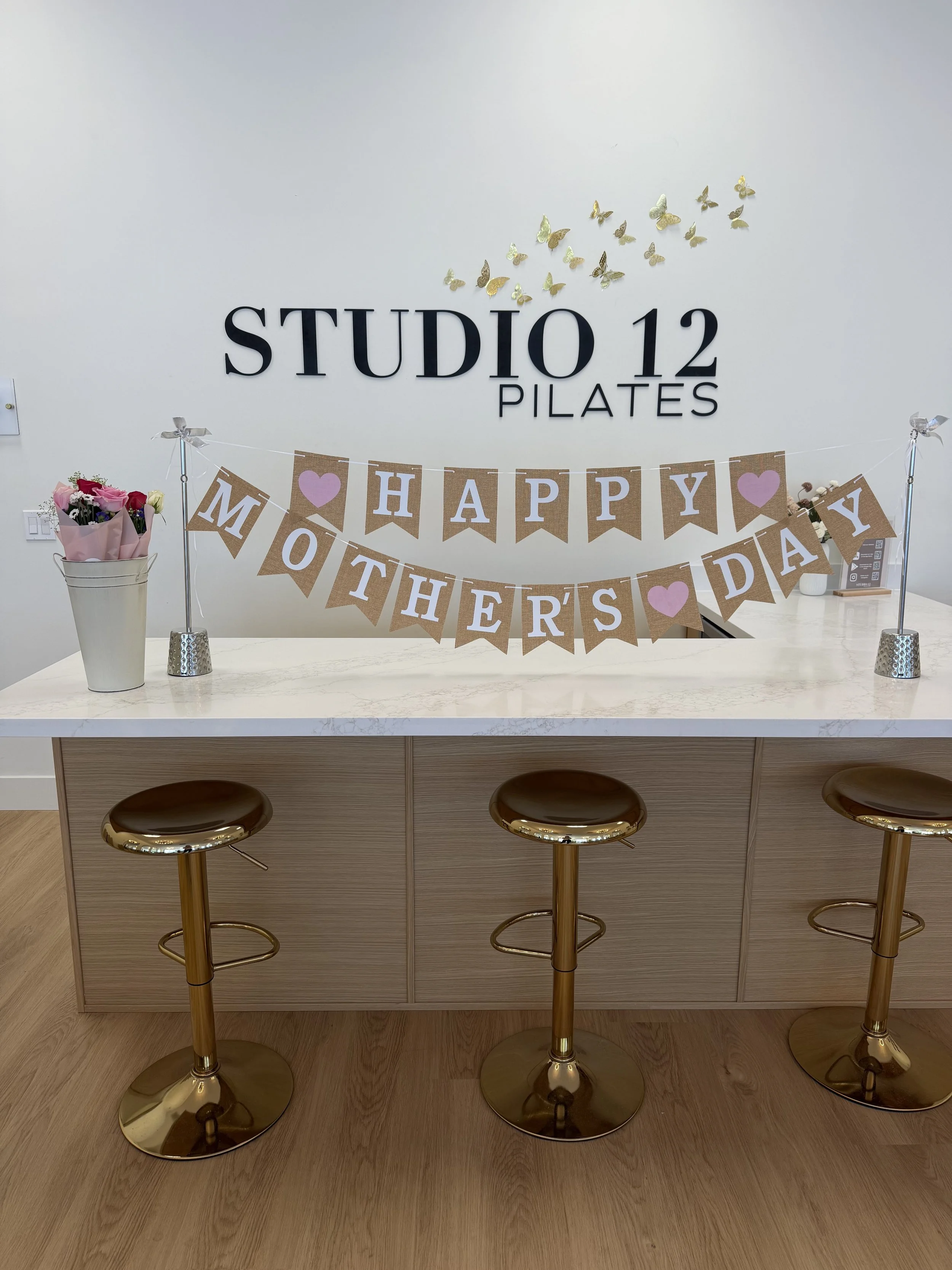 Reception area with a sign 'Studio 12 Pilates' on the wall, decorated with gold and silver butterfly wall decals, a banner that reads 'Happy Mother's Day' in brown paper with white letters and pink hearts, and a small table with a pink floral arrangement, three gold barstools, and a light-colored wooden floor.