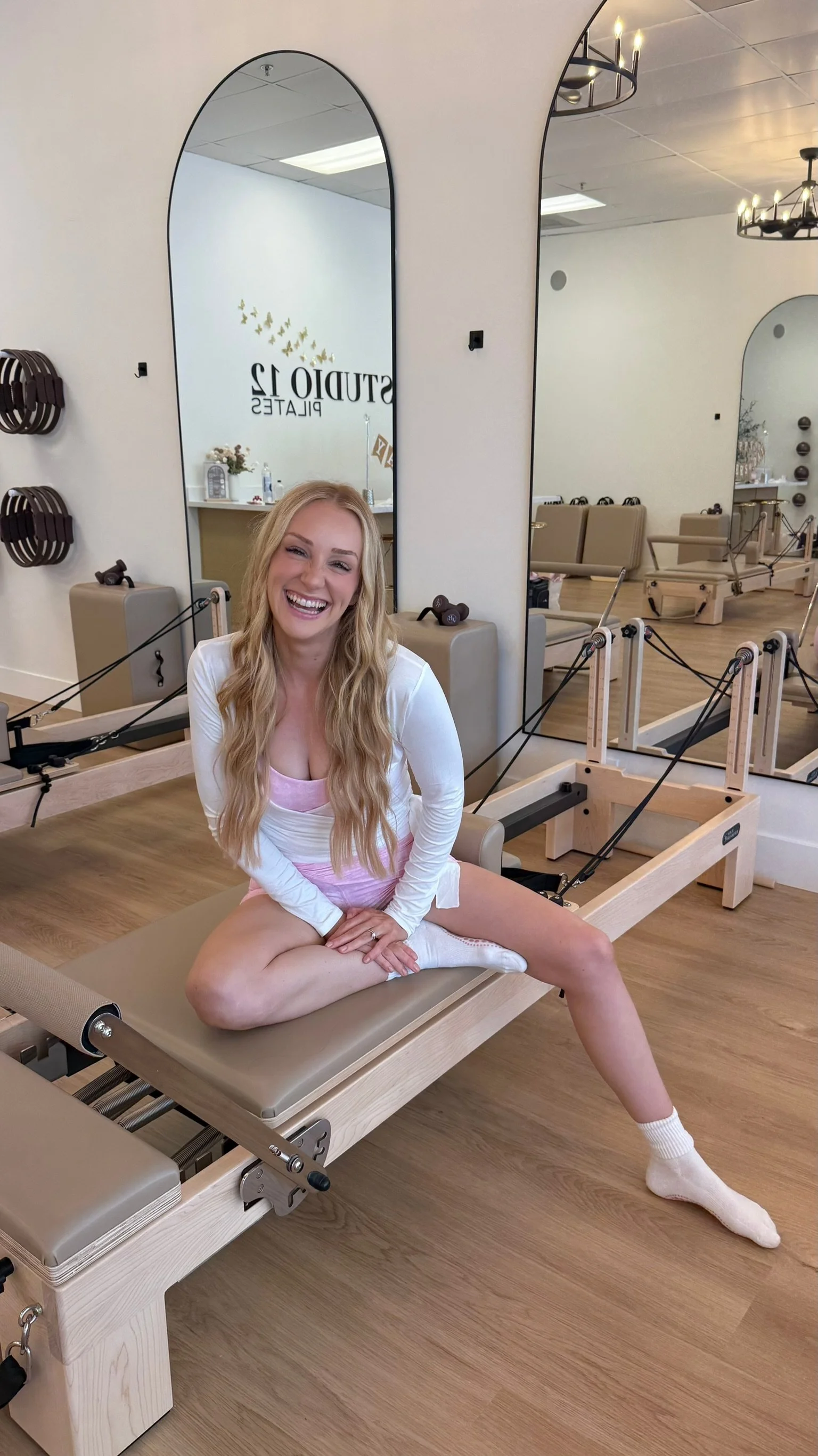 Happy woman sitting on Pilates reformer machine in a Pilates studio, smiling at the camera, wearing pink shorts and a white shirt.