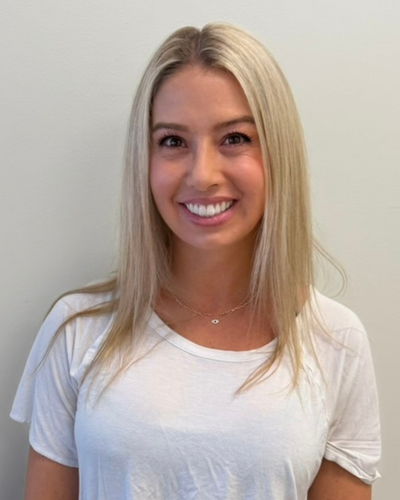 A young woman with long blonde hair, smiling, wearing a white t-shirt, standing against a plain light-colored wall.
