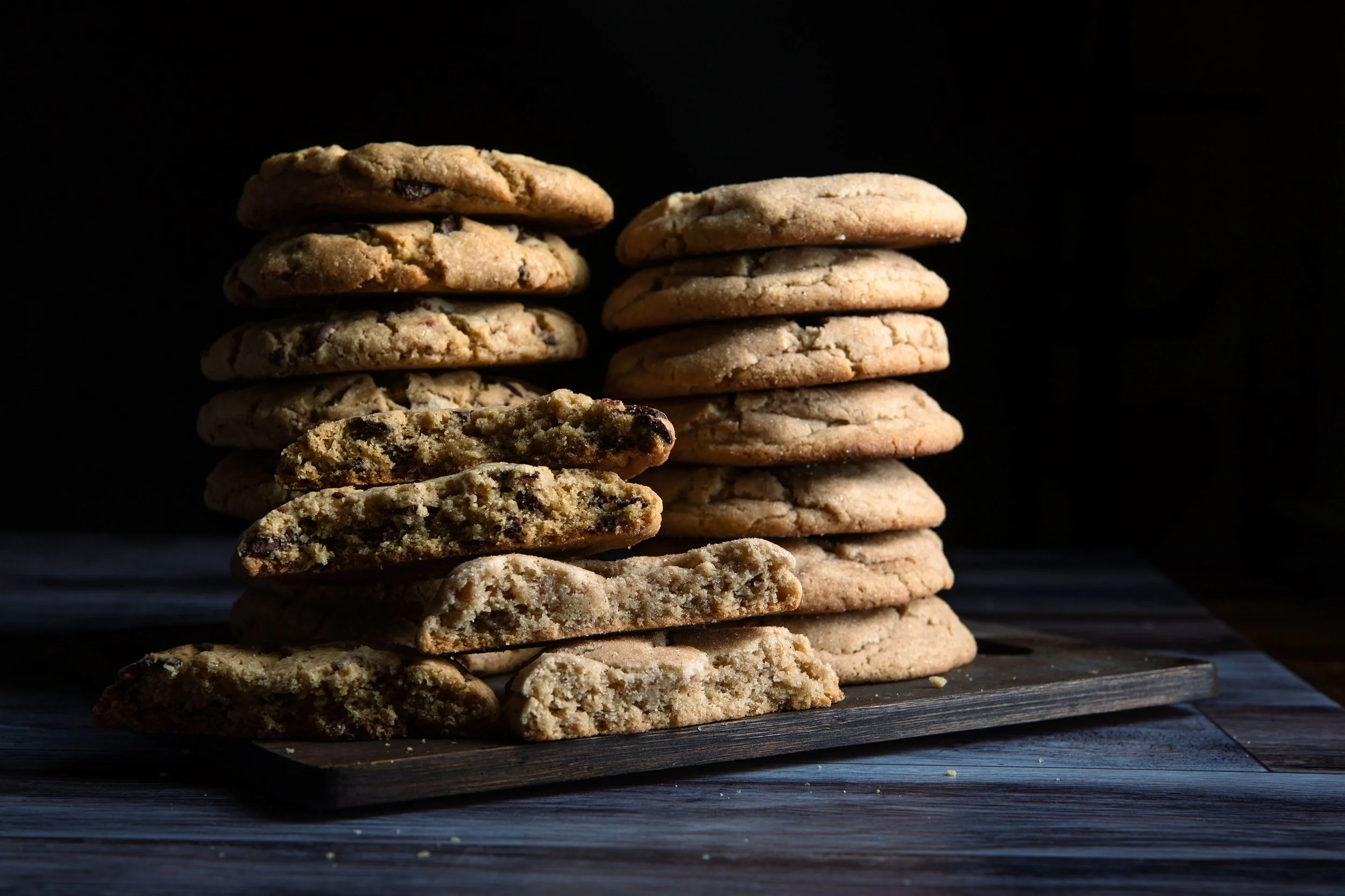 Stack of chocolate chip cookies on a dark wooden surface, with some broken cookie pieces in front.