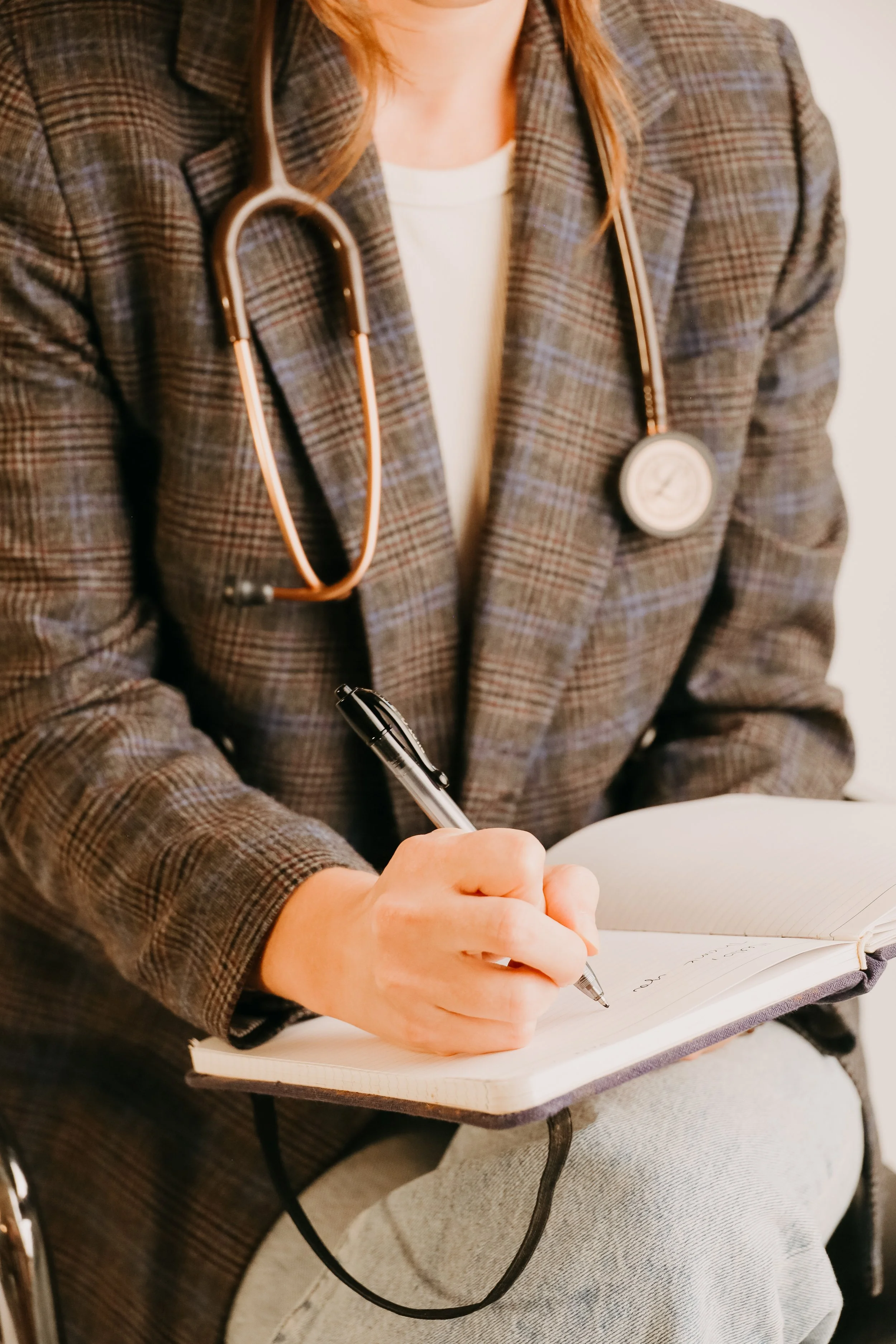 A woman in a plaid blazer with a stethoscope around her neck is writing in a notebook with a black pen.