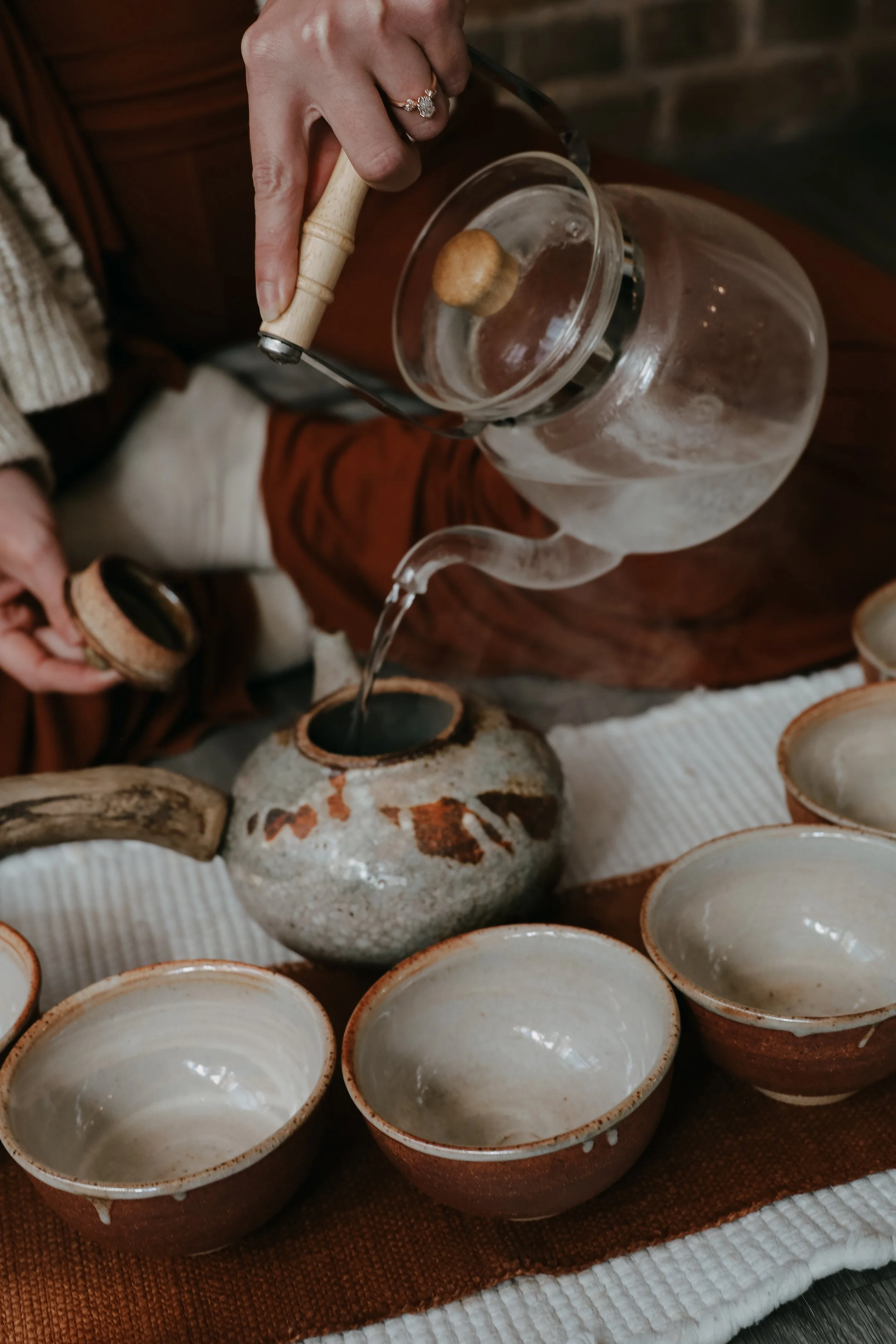 A person is pouring water from a clear glass kettle into a side-handle teapot on a table. It is a Tea Ceremony. The table has multiple empty ceramic bowls and a decorative ceramic vase, with a white cloth underneath.