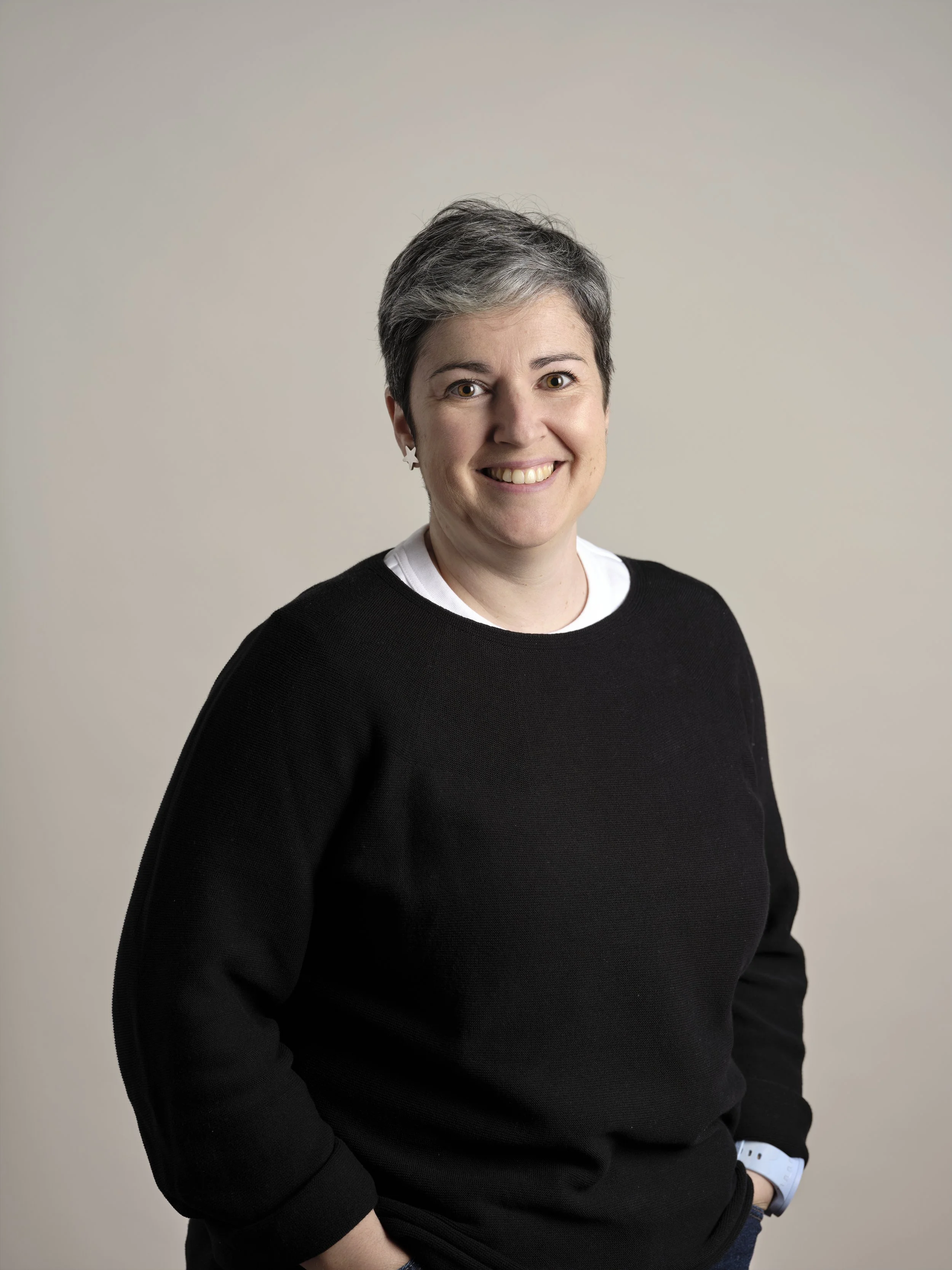 A woman with short gray hair smiling, wearing a black sweater over a white shirt, standing against a neutral background.