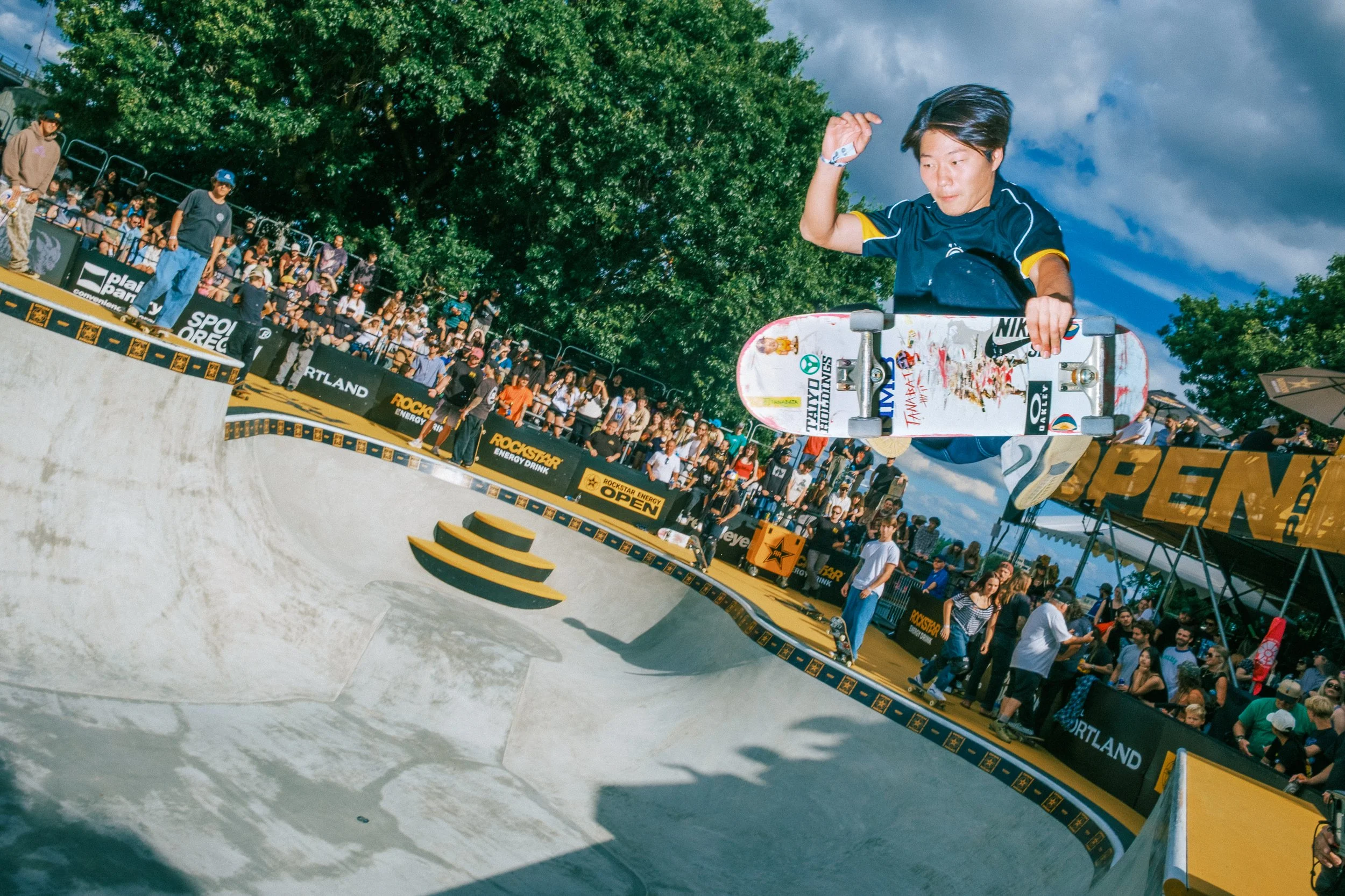 A skateboarder mid-air performing a trick in a skatepark, with a crowd of spectators watching.