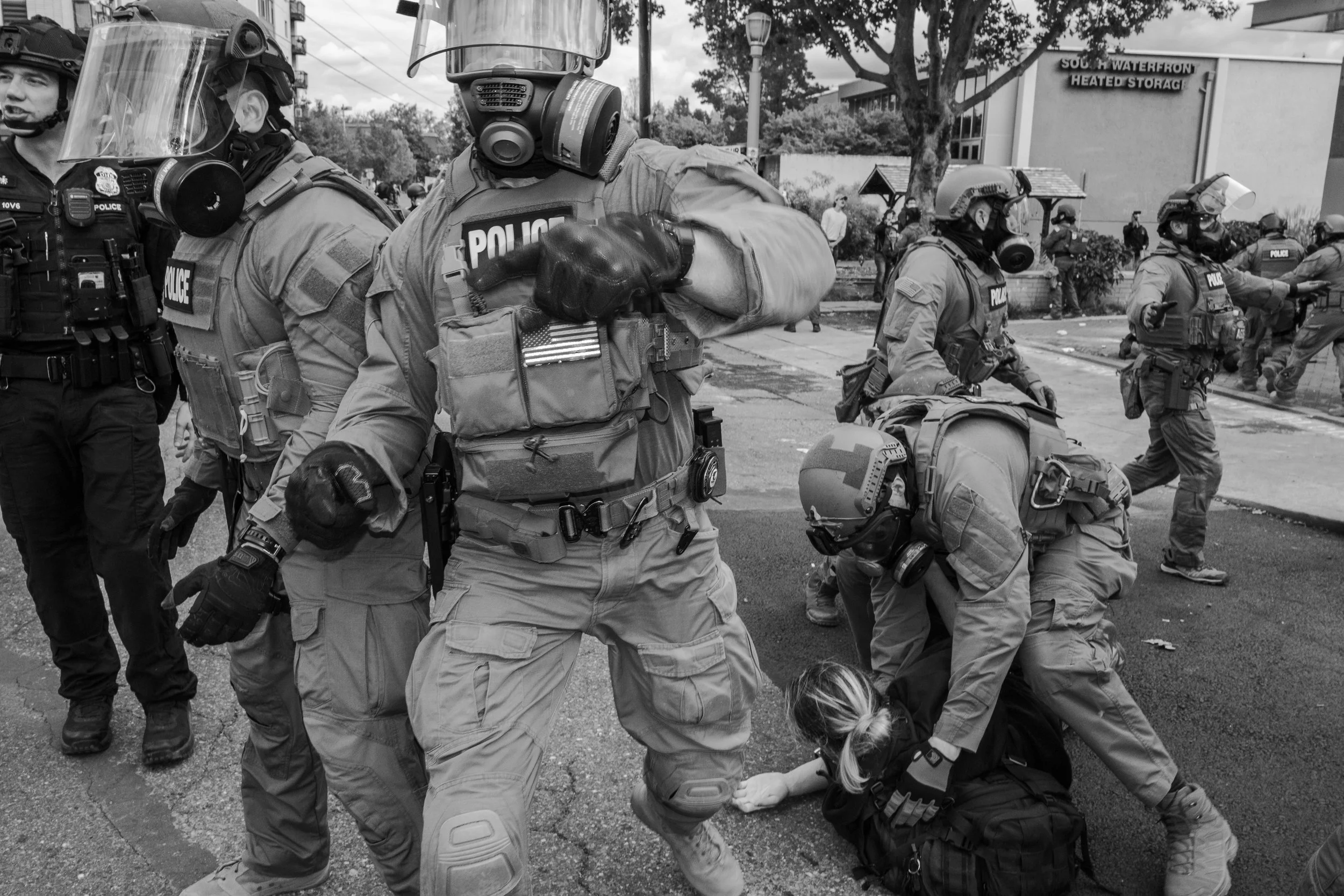 A group of police officers in tactical gear detain a person on the ground during a protest in an urban setting.