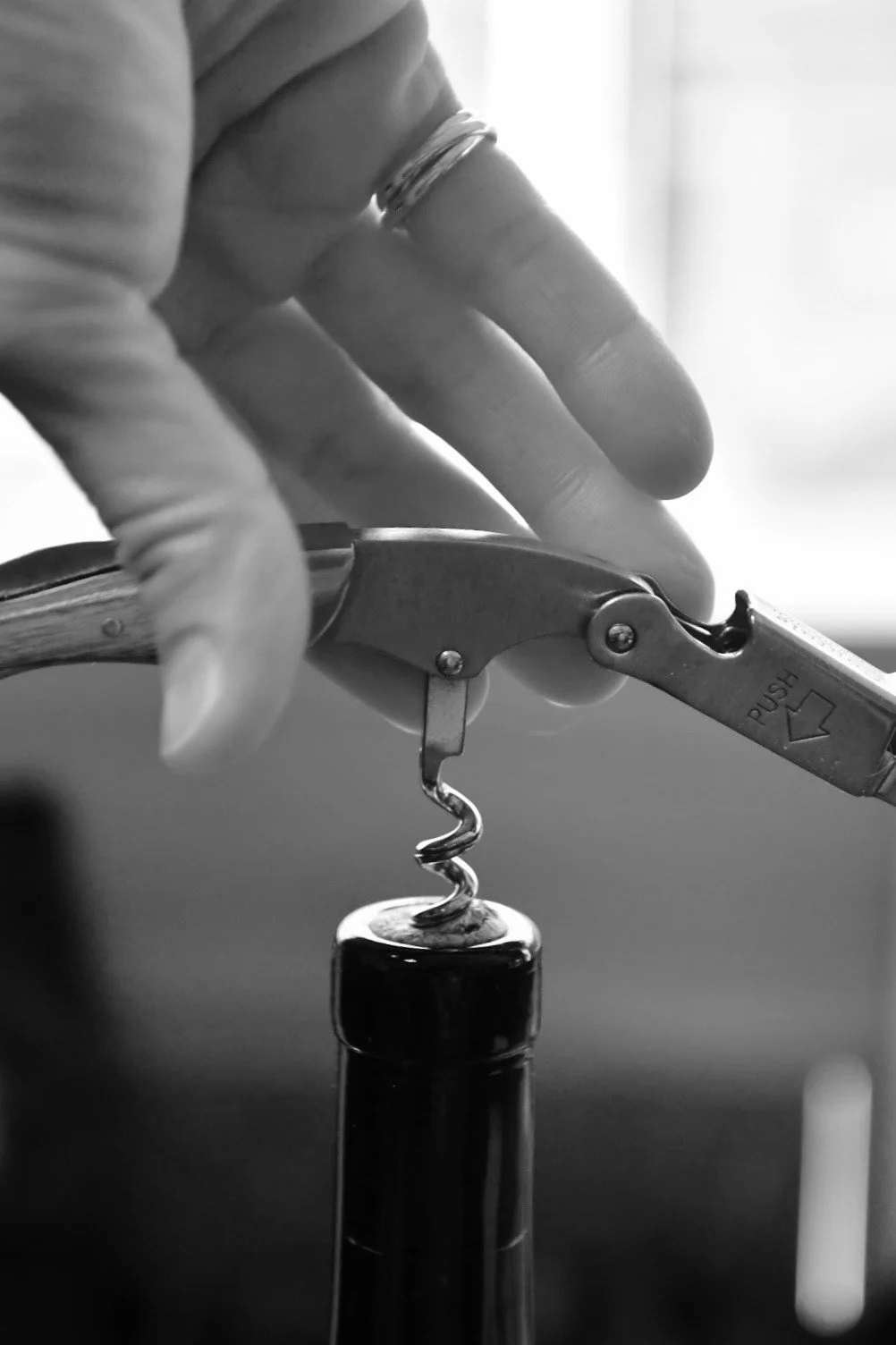 A black-and-white close-up of a hand opening a wine bottle with a corkscrew.