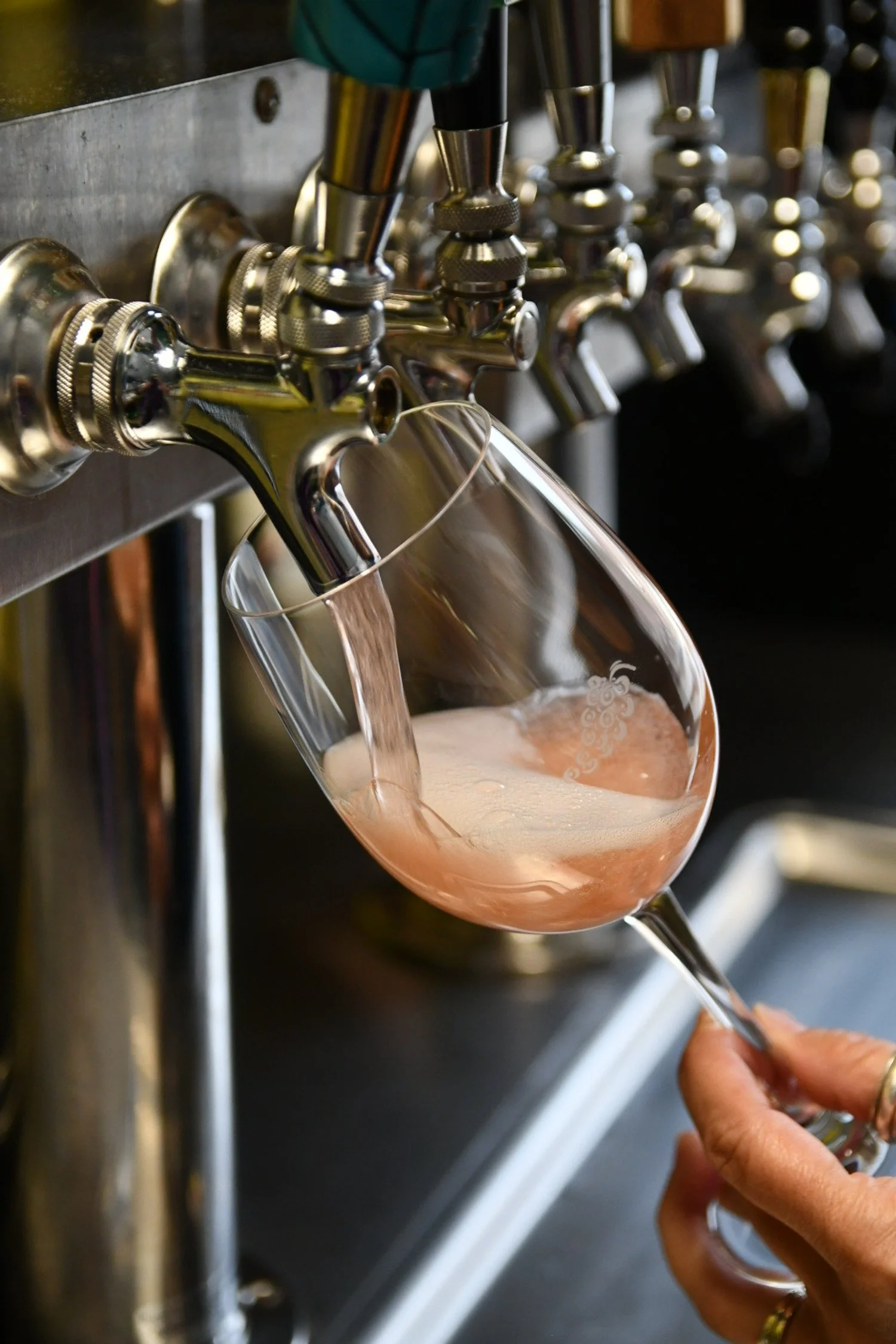 A person pours pink wine from a tap into a wine glass at a bar or winery.