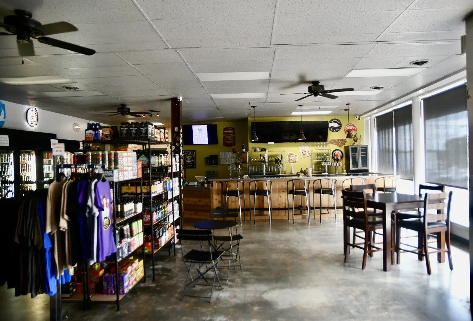 Interior of a brewery or bar with a wooden counter, bar stools, high tables with chairs, and display shelves with merchandise near large windows.