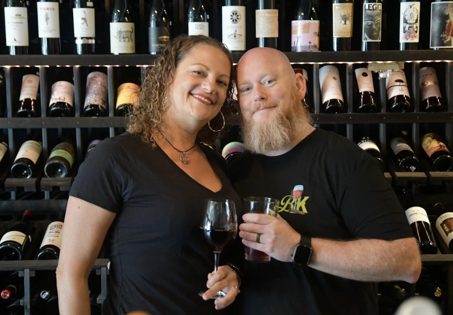 A woman and a man smiling and holding glasses of wine in front of a wine rack with bottles. The woman has curly hair, and the man has a beard and a watch on his left wrist.