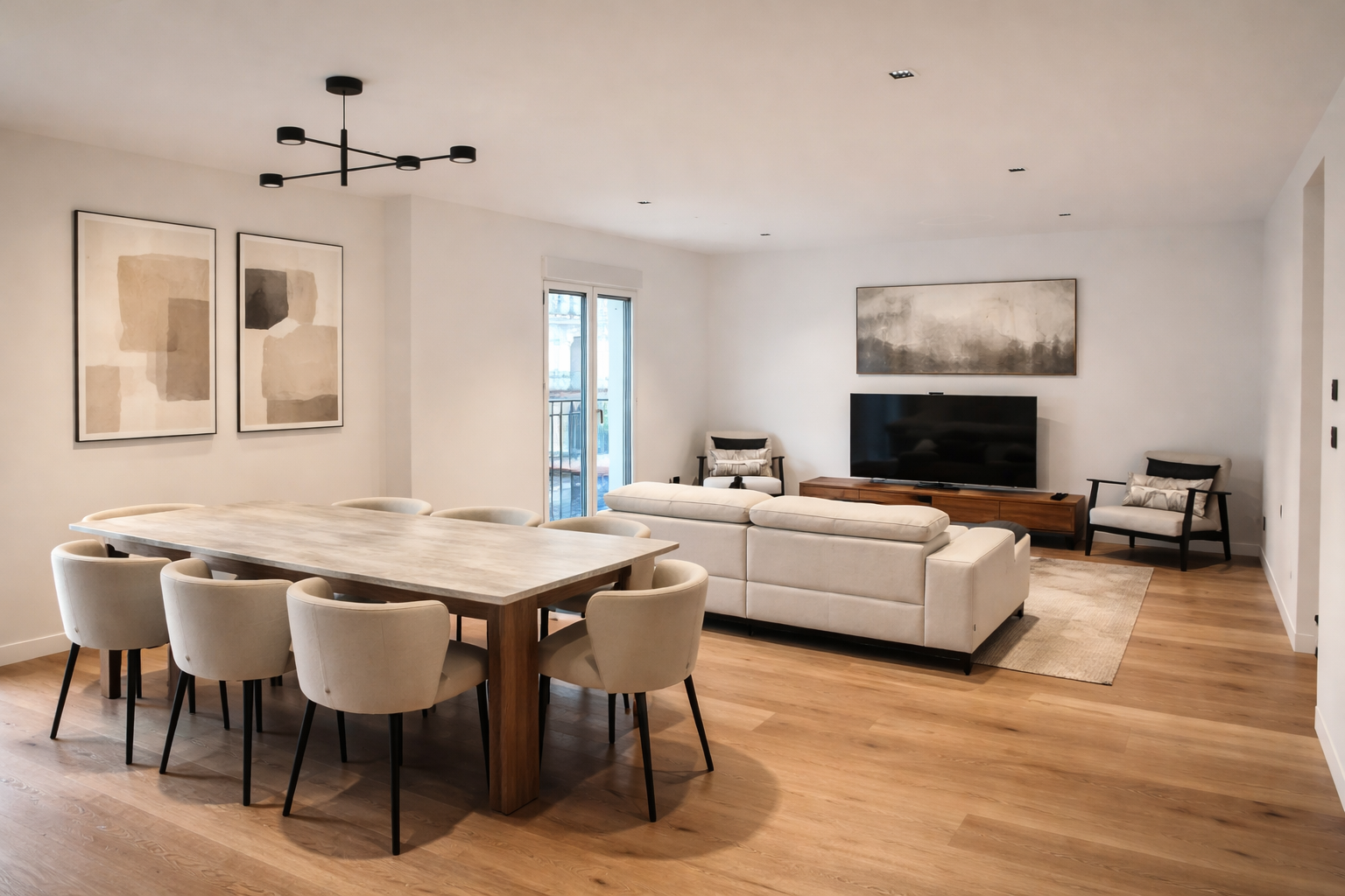 Living room and dining area with wooden floors, a large dining table surrounded by eight cream-colored chairs, a white sectional sofa facing a television, and two black armchairs near the window. Modern abstract wall art and a black ceiling fixture a