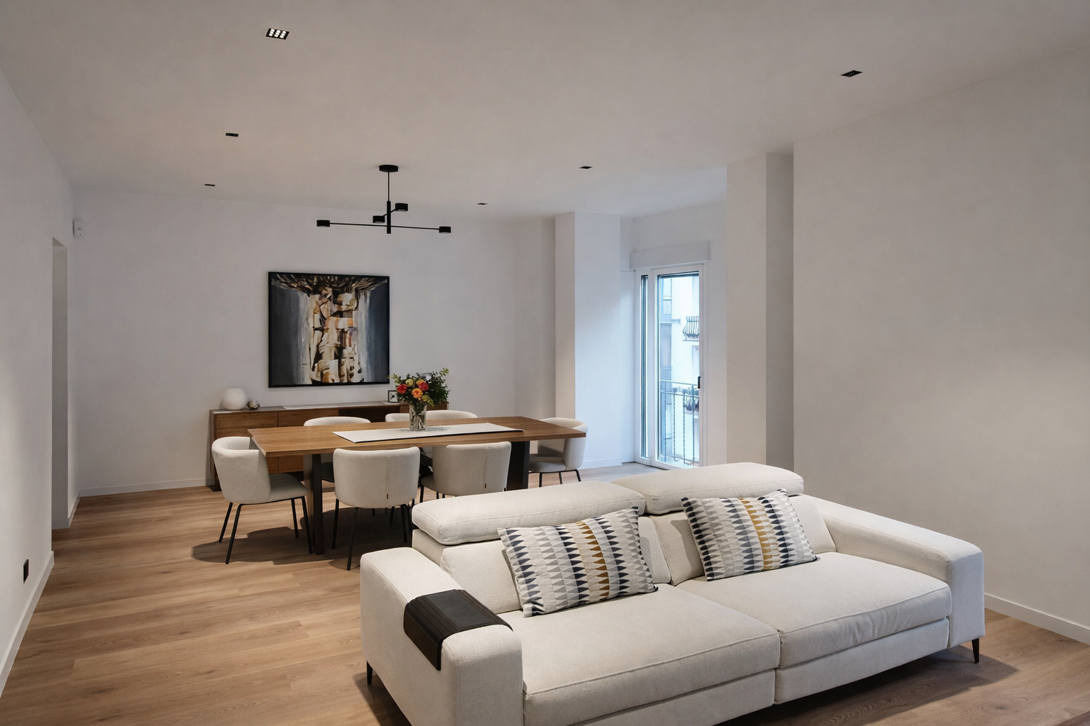 Modern living and dining room with white walls, light wood flooring, a white sofa with patterned pillows, a wooden dining table with seven cream chairs, a sideboard with a vase, a painting on the wall, a black ceiling chandelier, and a glass door lea