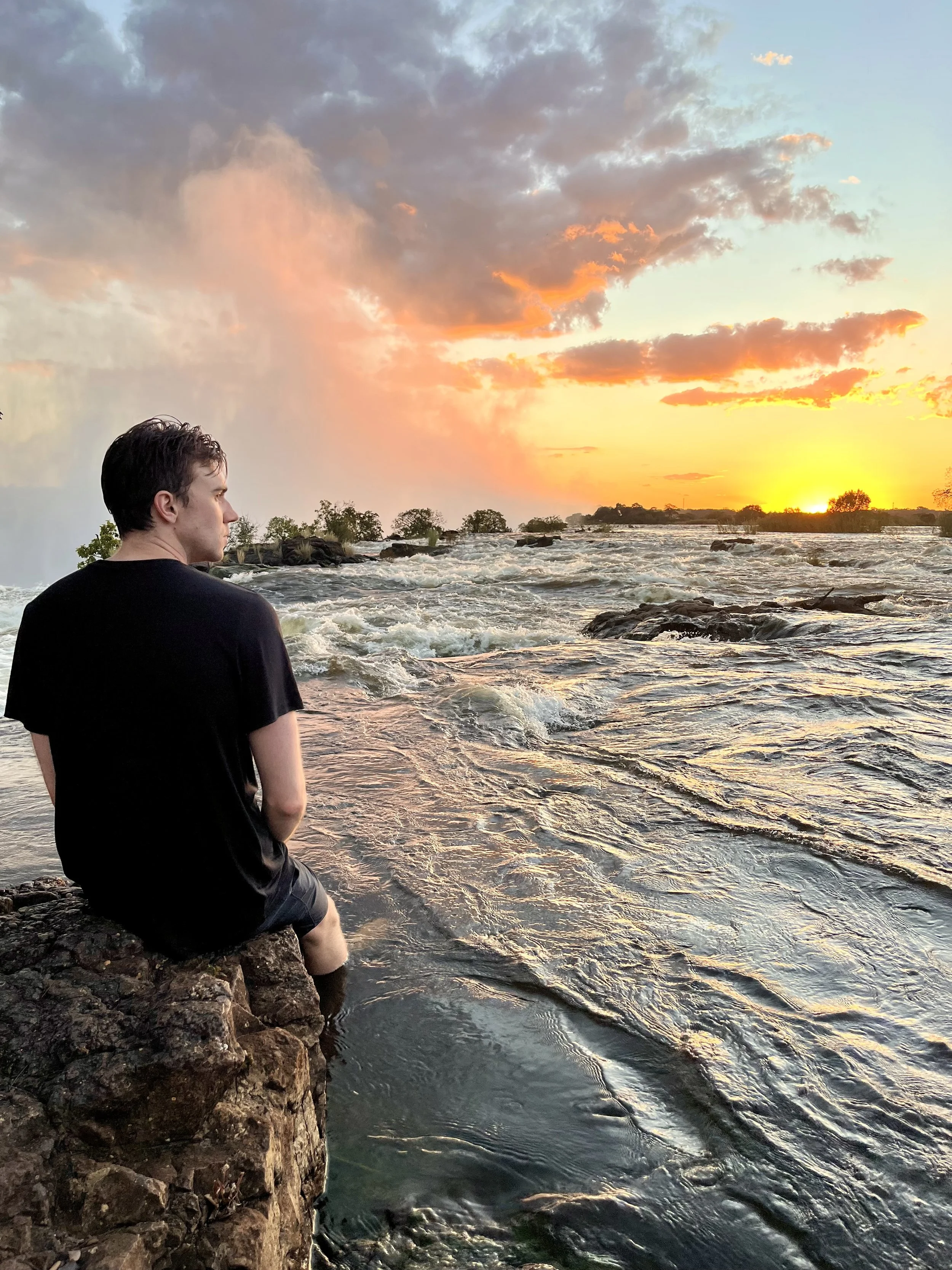 A person sitting on rocks at the edge of a river during sunset, with trees in the distance and partly cloudy sky.