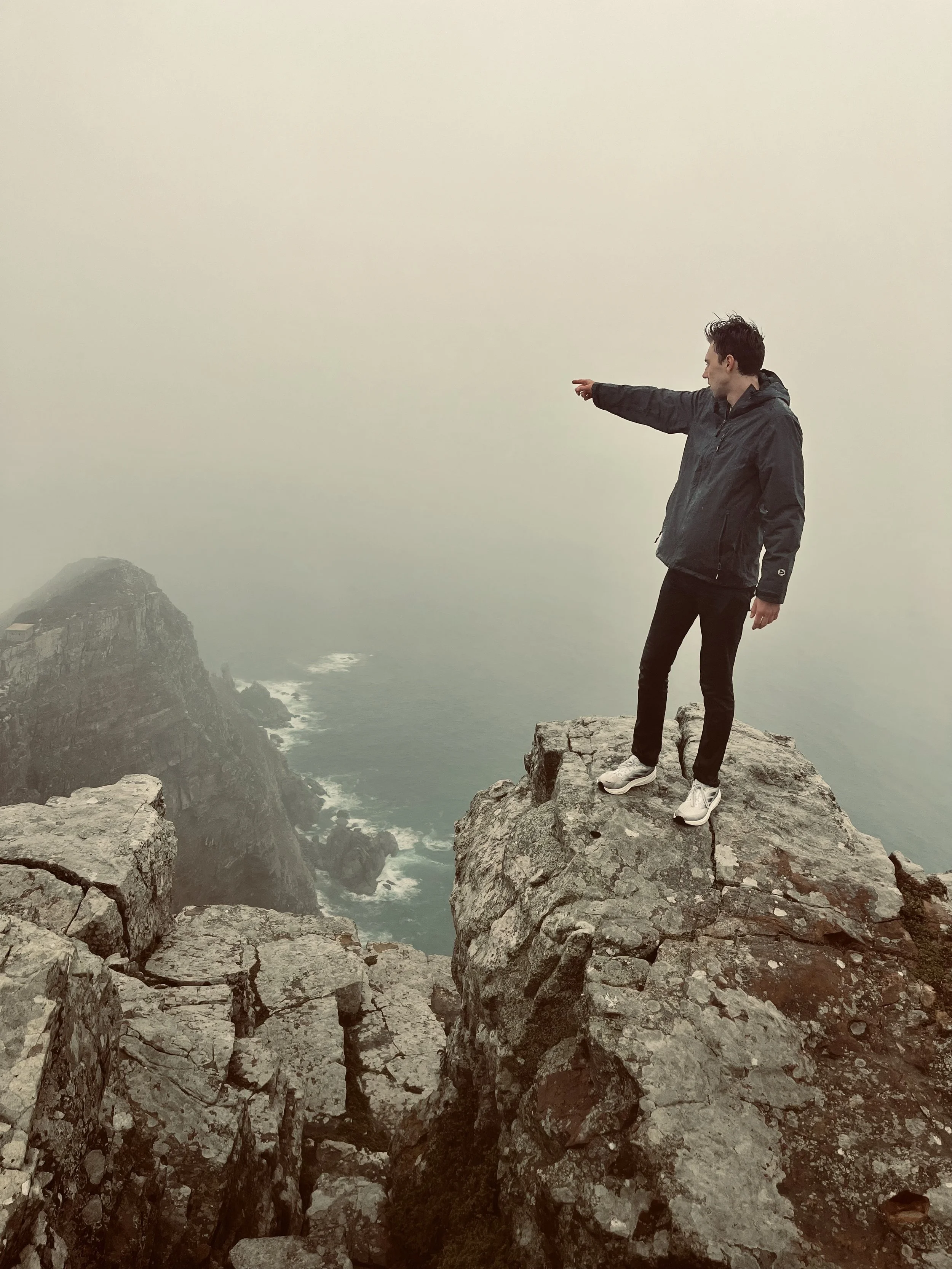 A person standing on the edge of a rocky cliff overlooking a foggy ocean, pointing towards the distance.