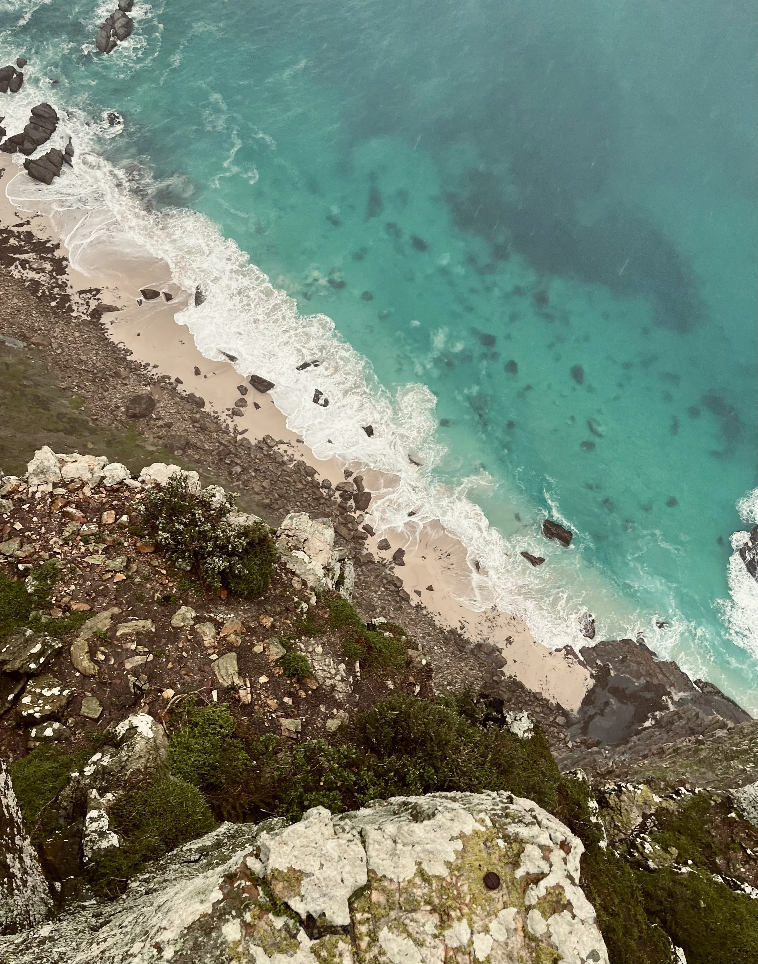 Aerial view of a rocky coastline with waves crashing onto a sandy beach and turquoise ocean waters.
