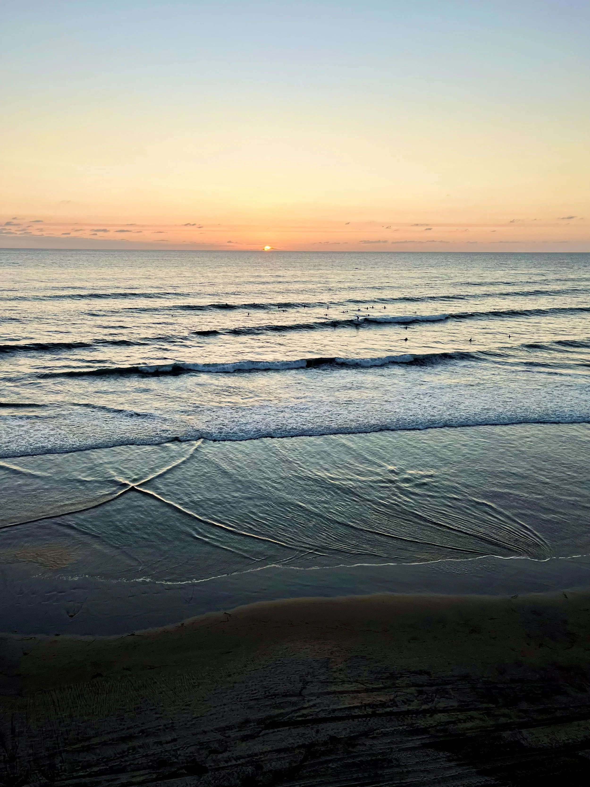 Sunset over the ocean with gentle waves and a few birds swimming. The sky displays soft pastel colors.