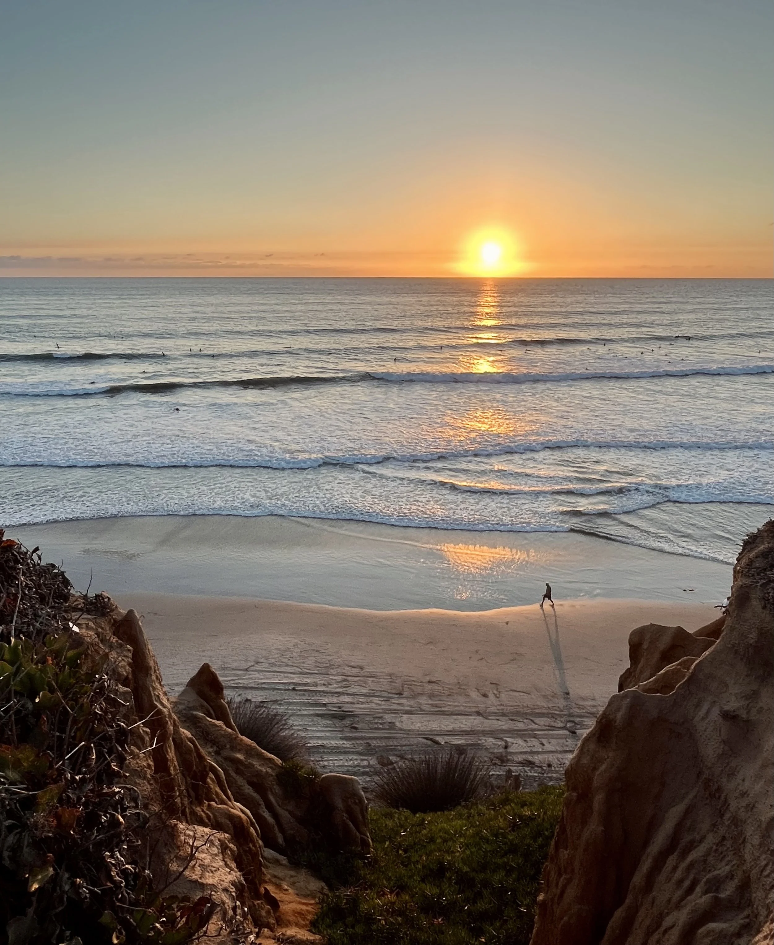 Sunset over the ocean with waves, a person walking on the beach, rocks and plants in the foreground