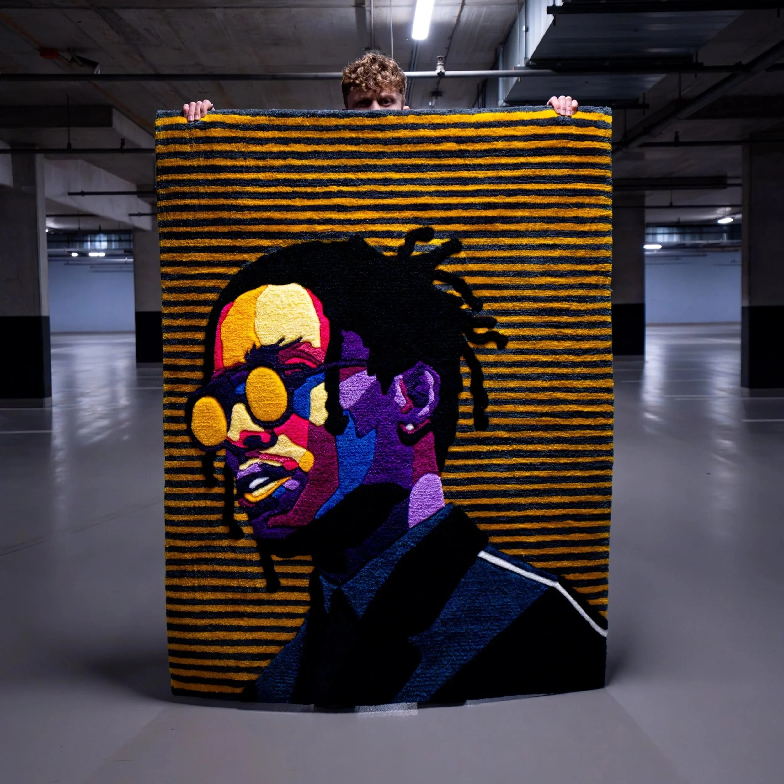An art piece or rug featuring a colorful portrait of a man with dreadlocks and sunglasses, set against a background of horizontal black and yellow stripes.