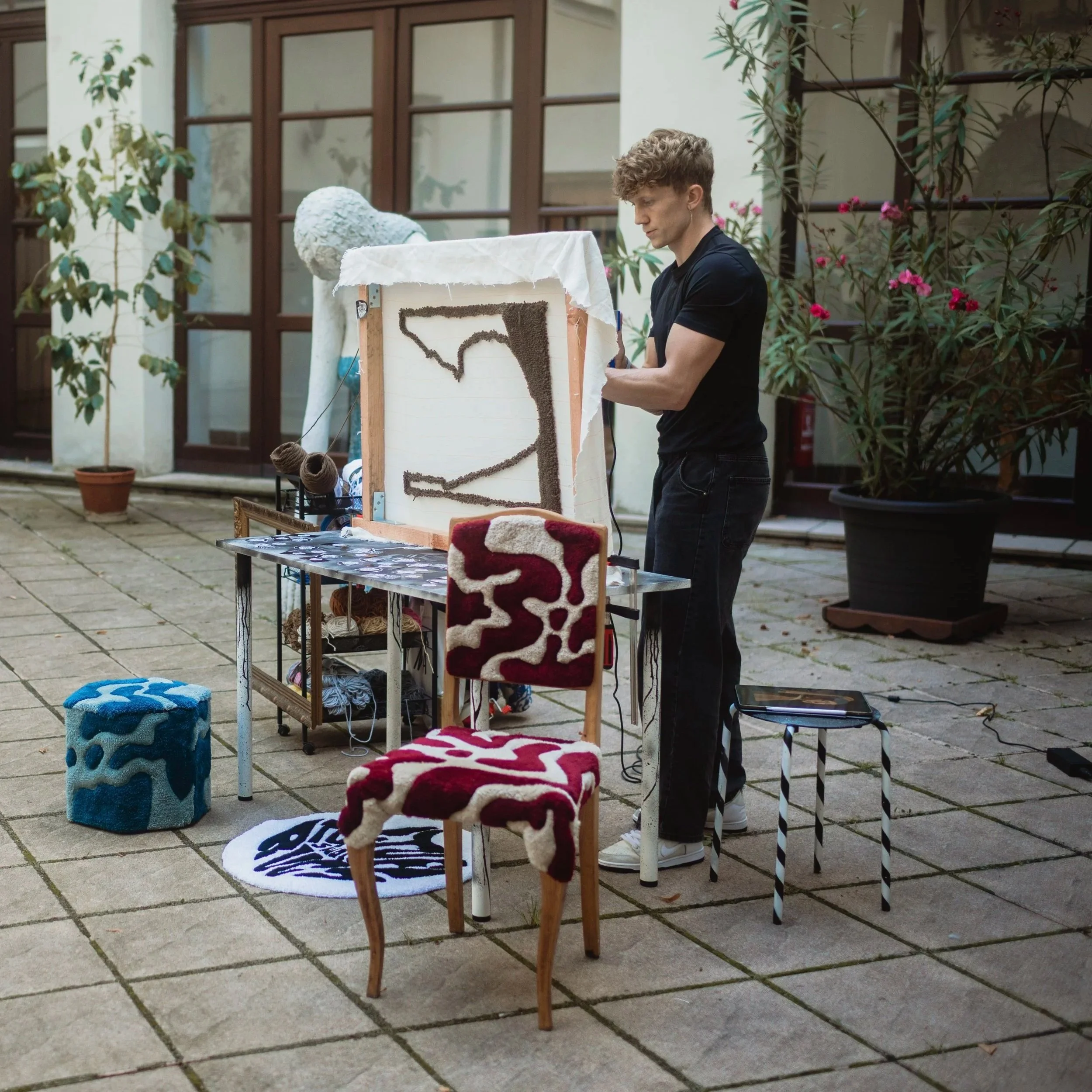 A man creating textile art on a small outdoor setup with chairs, a table, and potted plants nearby.