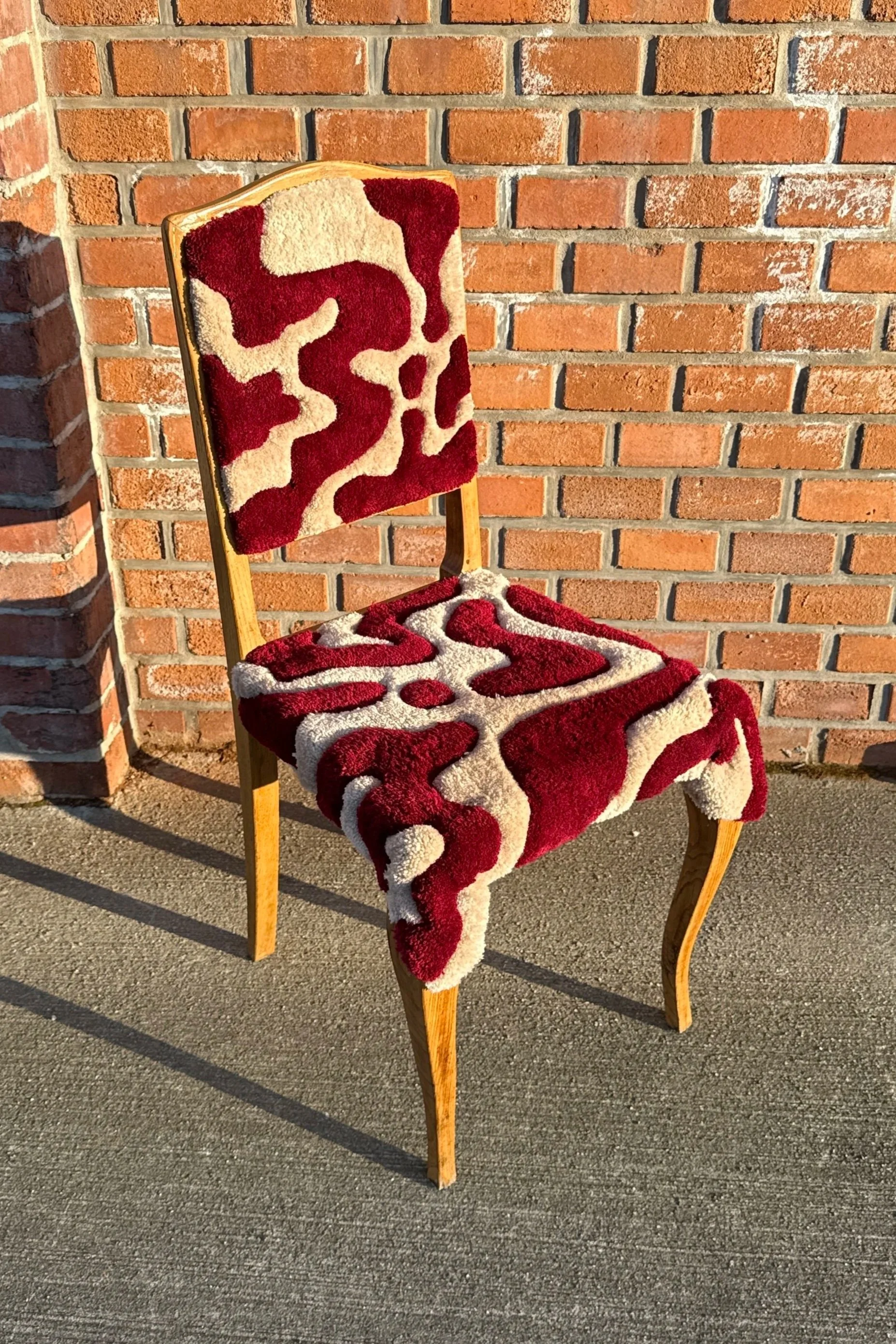 A vintage wooden chair with a patterned, plush red and beige cushion on the seat and backrest, positioned against a brick wall outside.