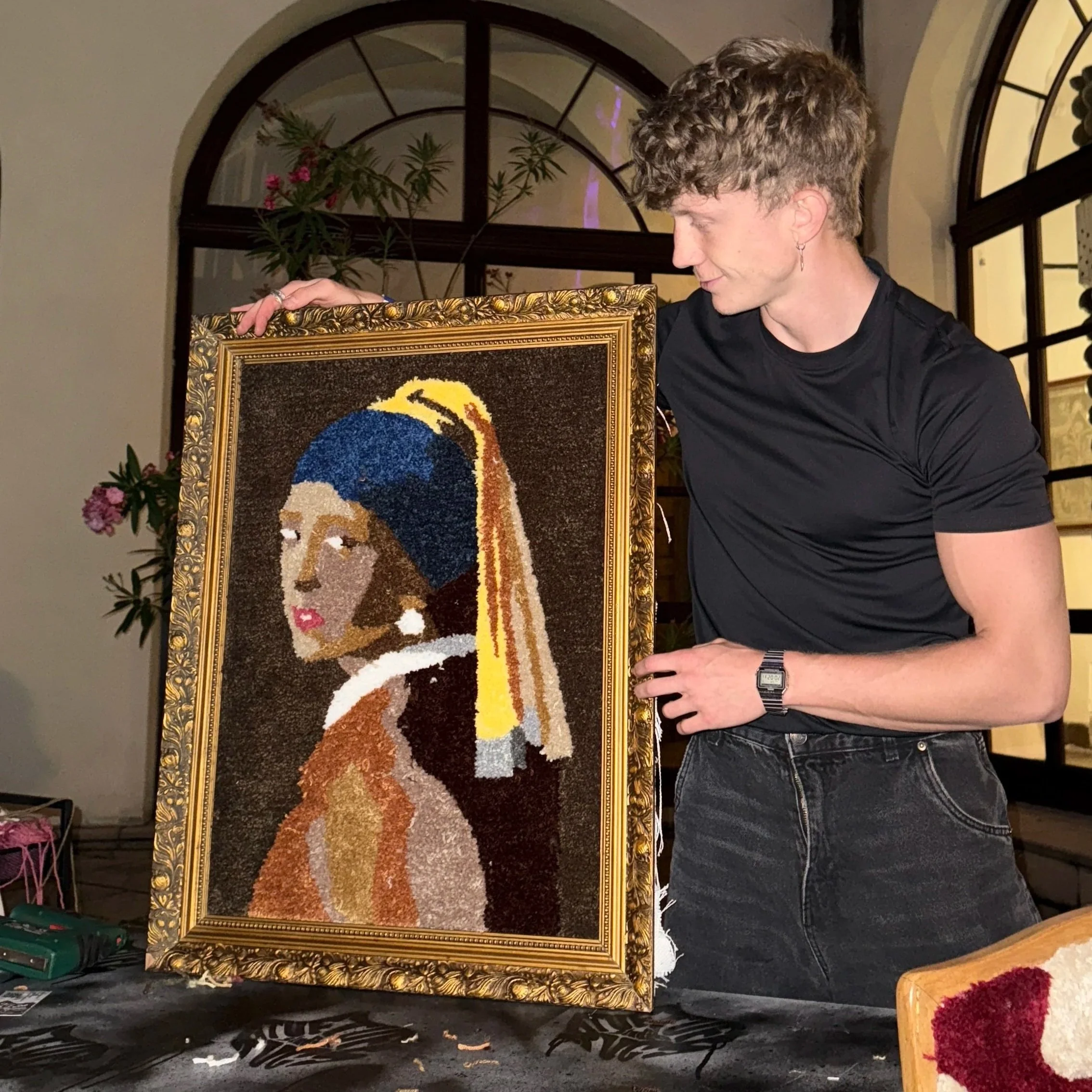 A young man with curly hair is holding a framed artwork of a woman with a blue headscarf and white earring, made from colored materials, in a room with large windows and indoor plants.