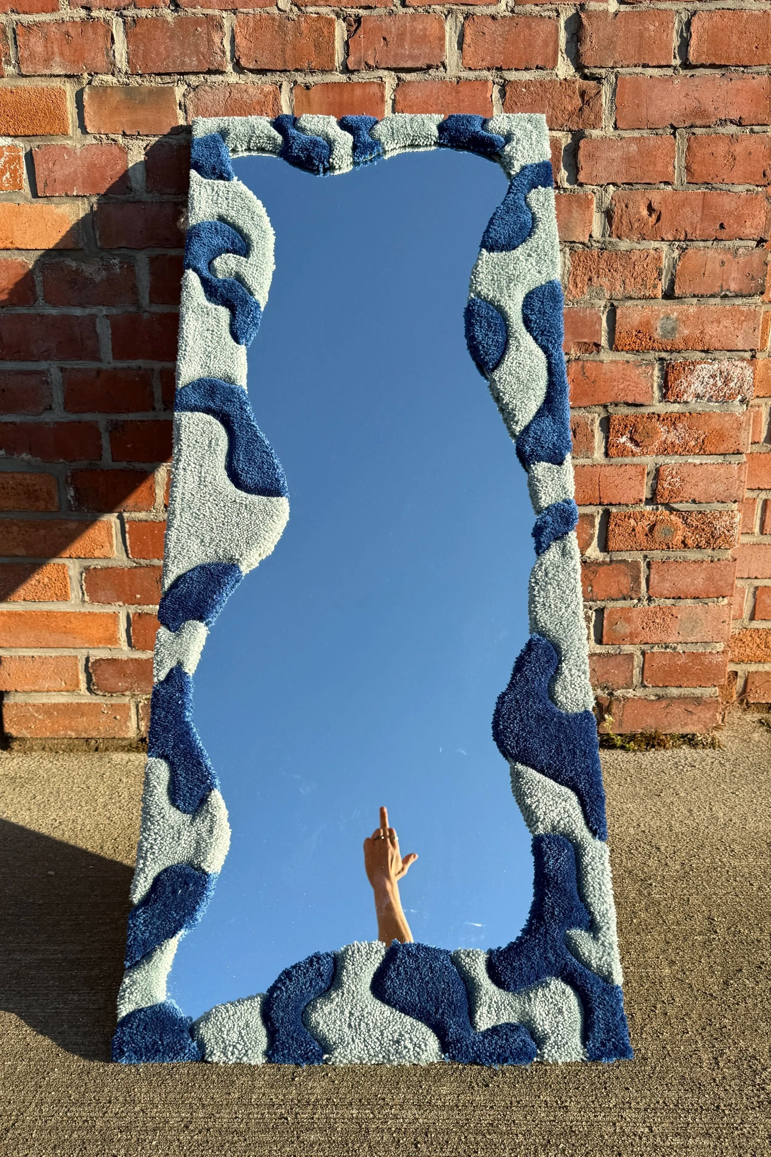 A decorative mirror with a blue and white textured frame, leaning against a brick wall, reflecting a hand making a gesture and a clear blue sky.