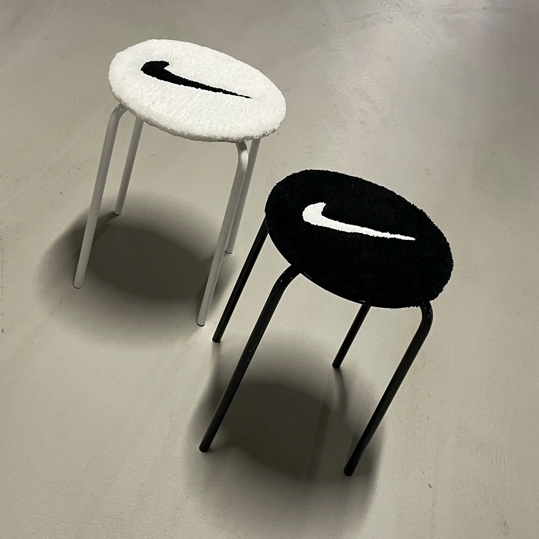 Two small stools with Nike logos on the seats, one with a white seat and black logo, the other with a black seat and white logo, on a light-colored floor.