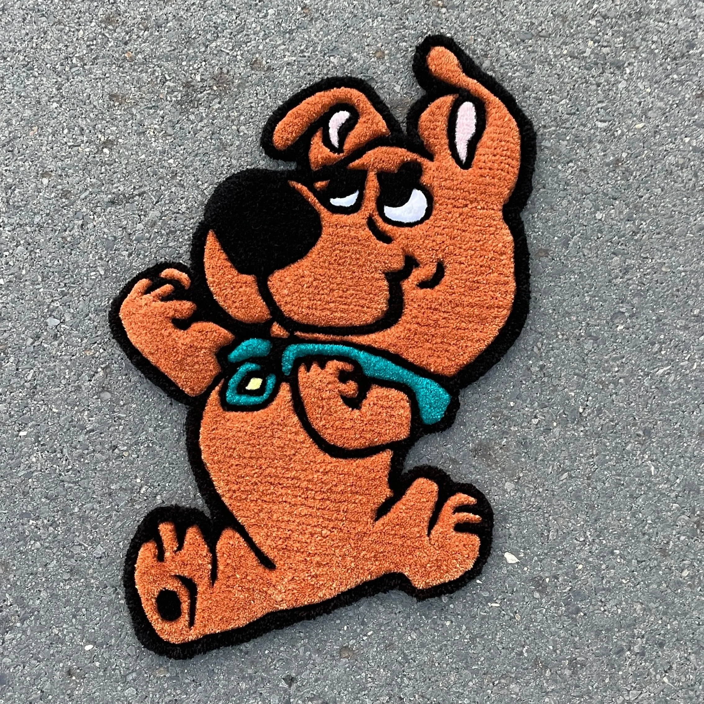 Colorful embroidered patch of Scooby-Doo, a cartoon dog, with one eye open, a blue collar, and a playful expression, on a gray surface.