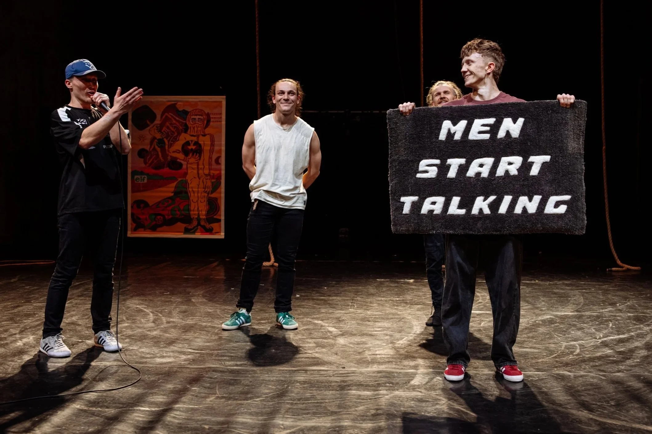 Four people standing on a stage. One person is holding a microphone and speaking, while another holds a sign that reads 'MEN START TALKING'. The background has a poster and dark curtains.
