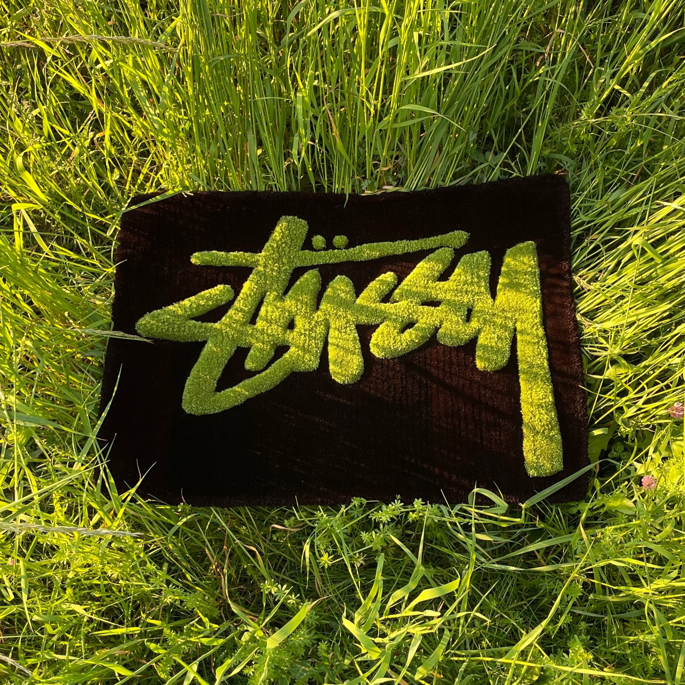 A black patch with a yellowish-green embroidered design of text, placed on green grass and plants