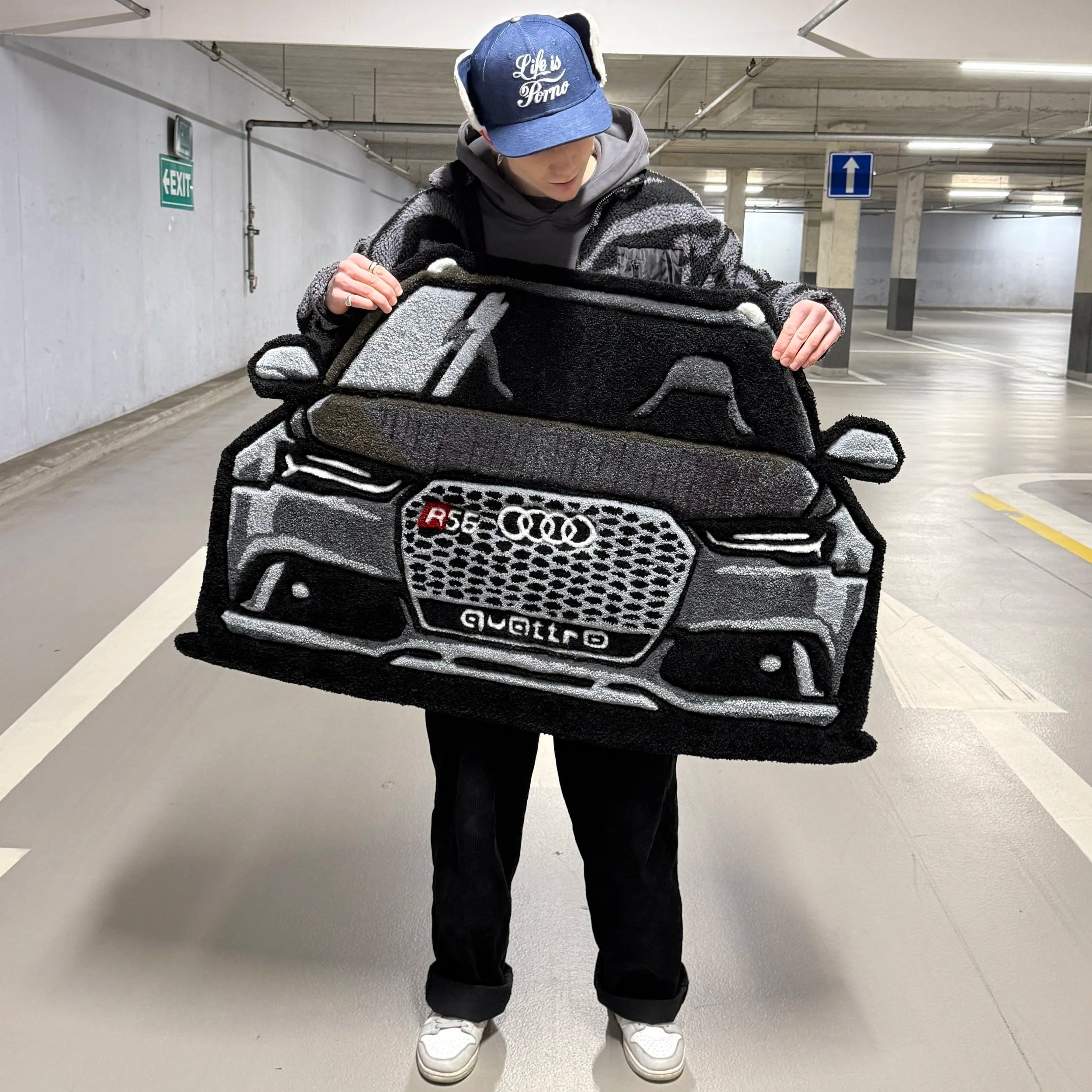 Person holding a rug designed to look like a black Audi car, inside a parking garage.