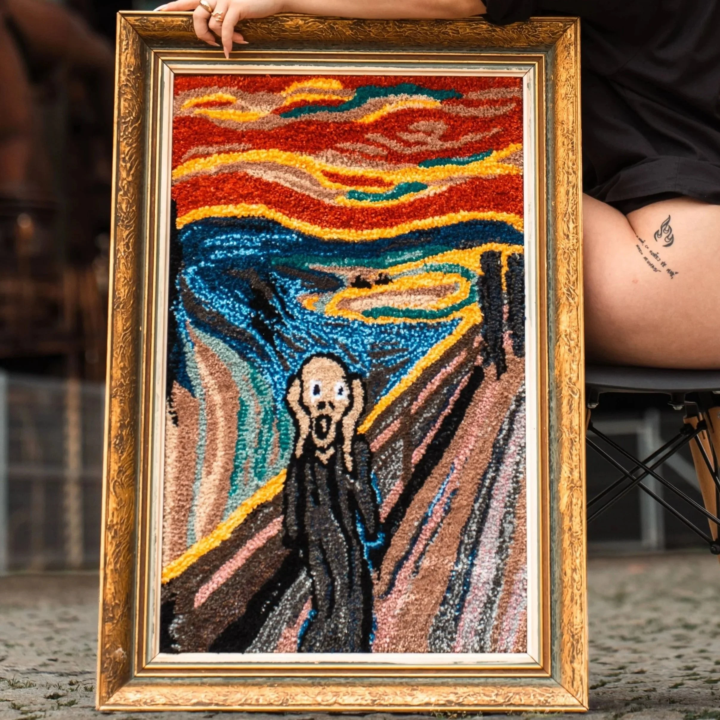 A woman with red hair wearing black sunglasses and a black outfit, sitting with her arm resting on a framed artwork of "The Scream" made from a colorful mosaic, outdoors in an industrial setting.