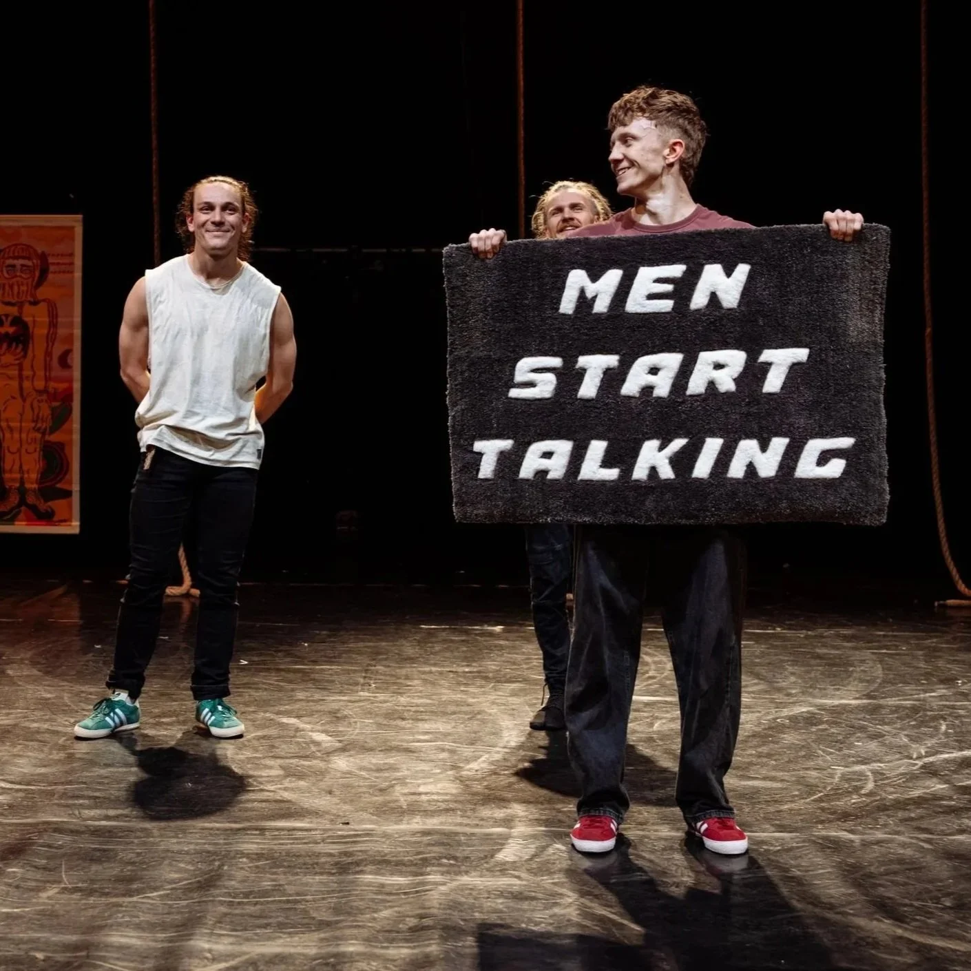 Four people standing on a stage. One person is holding a microphone and speaking, while another holds a sign that reads 'MEN START TALKING'. The background has a poster and dark curtains.